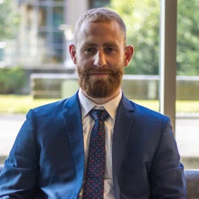 Man with beard in a dark blue suit, white shirt, and patterned tie sitting indoors near a large window with greenery outside.