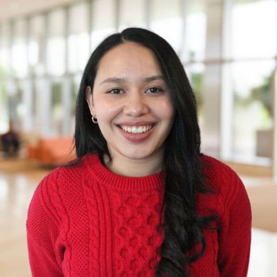 A woman smiling and wearing a red sweater in a bright room with large windows.