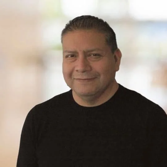 A man with short dark hair, wearing a black shirt, smiling against a blurred background.