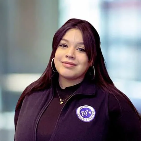 A woman with long dark red hair and hoop earrings, wearing a black shirt and a black zip-up jacket with a BJ's Wholesale Club logo. She is smiling slightly and standing indoors with a blurred background.