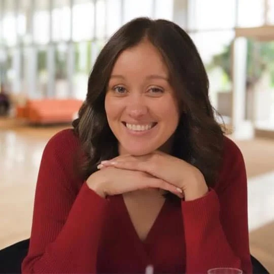 Woman with shoulder-length dark hair, smiling, wearing a red sweater, sitting at a table in a bright indoor setting.