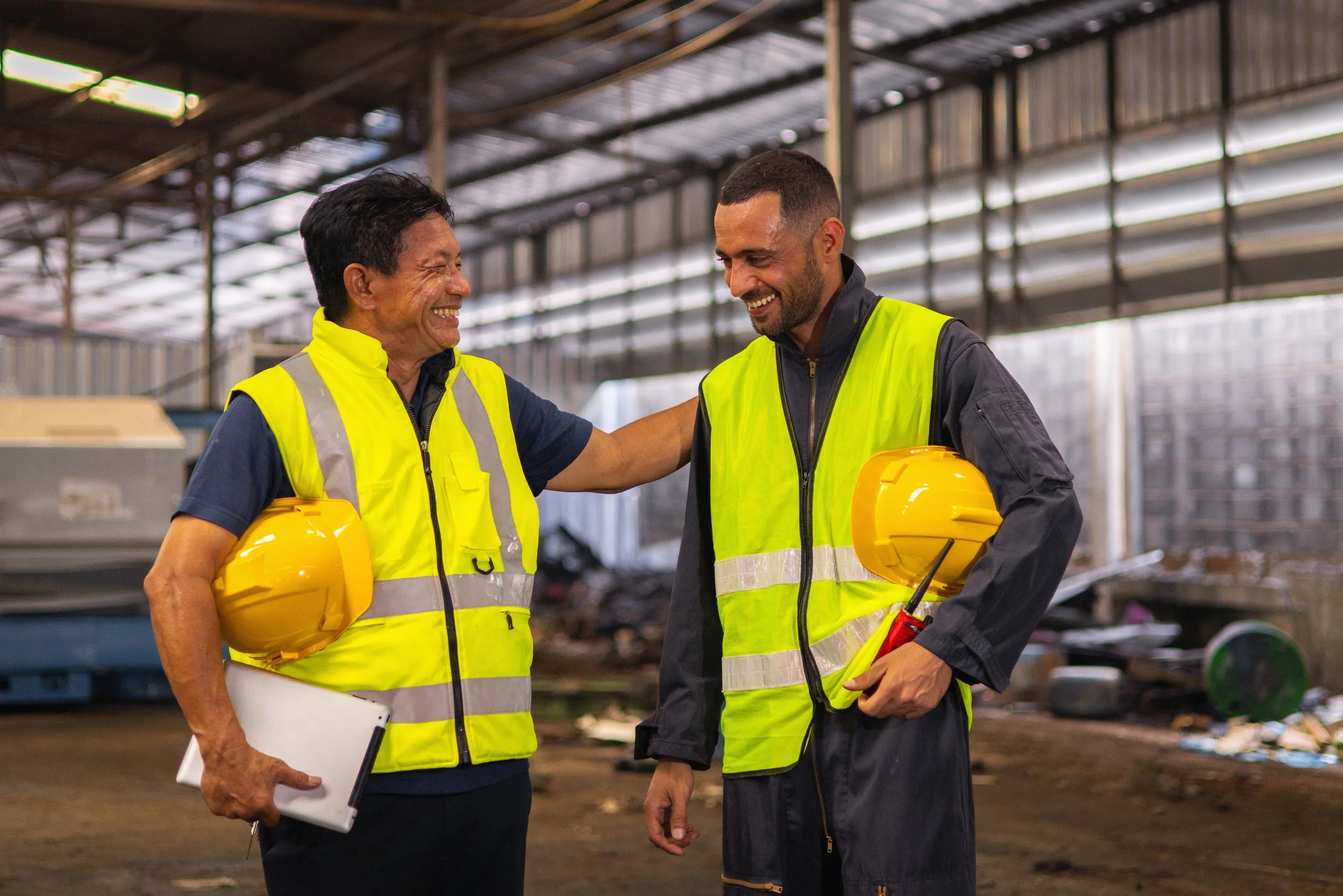 Two construction workers, a woman and a man, wearing high-visibility safety vests and yellow hard hats, are smiling and engaging in conversation inside a construction site with metal framework and building materials around them.