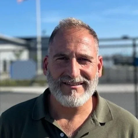 A smiling middle-aged man with a beard and short gray hair standing outdoors on a sunny day.