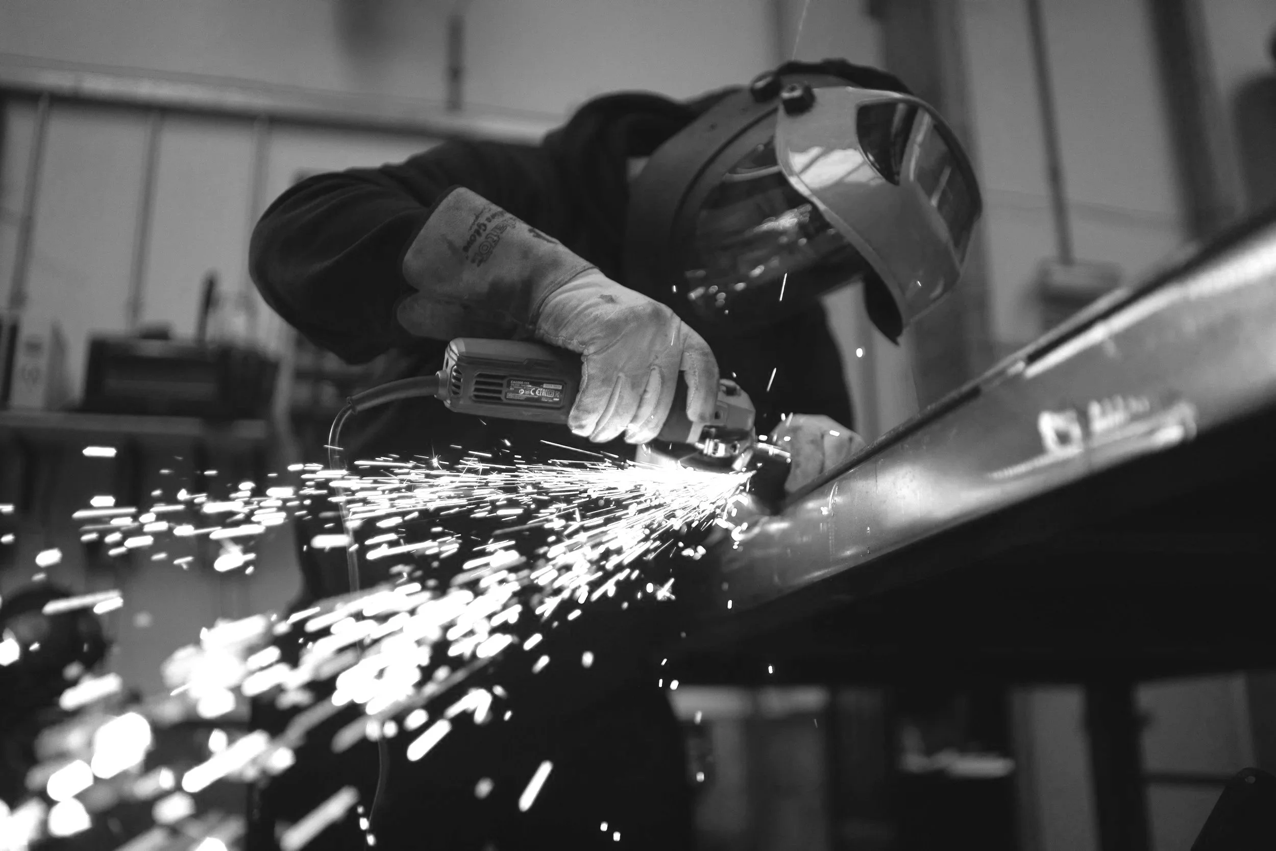 A person welding metal in a workshop, wearing protective gear including gloves and a welding mask, with sparks flying from the welding process.