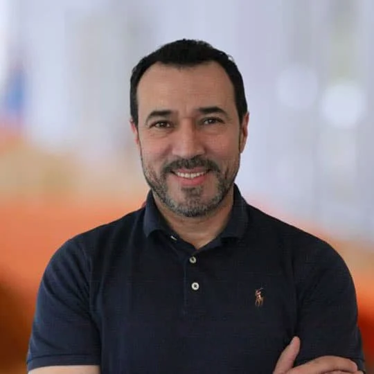 A man with dark hair, beard, and mustache, smiling while wearing a navy polo shirt with a small emblem, standing against a blurred background.