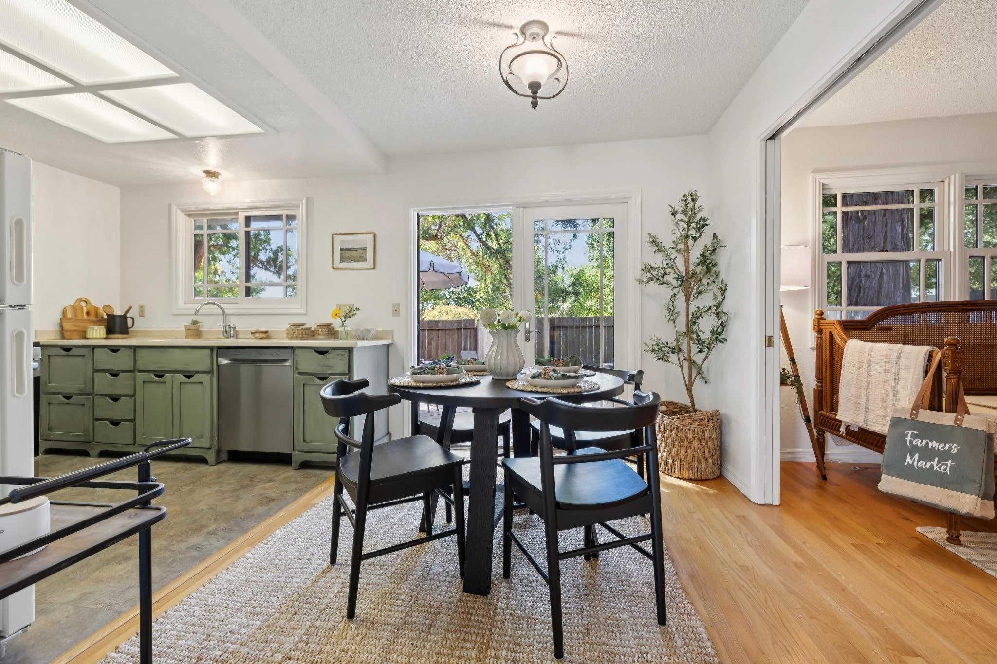 A dining area adjacent to a kitchen with a round black table set with plates and napkins, surrounded by black chairs. The room features large windows and a sliding glass door revealing an outdoor view with trees. A vase with white flowers and other d