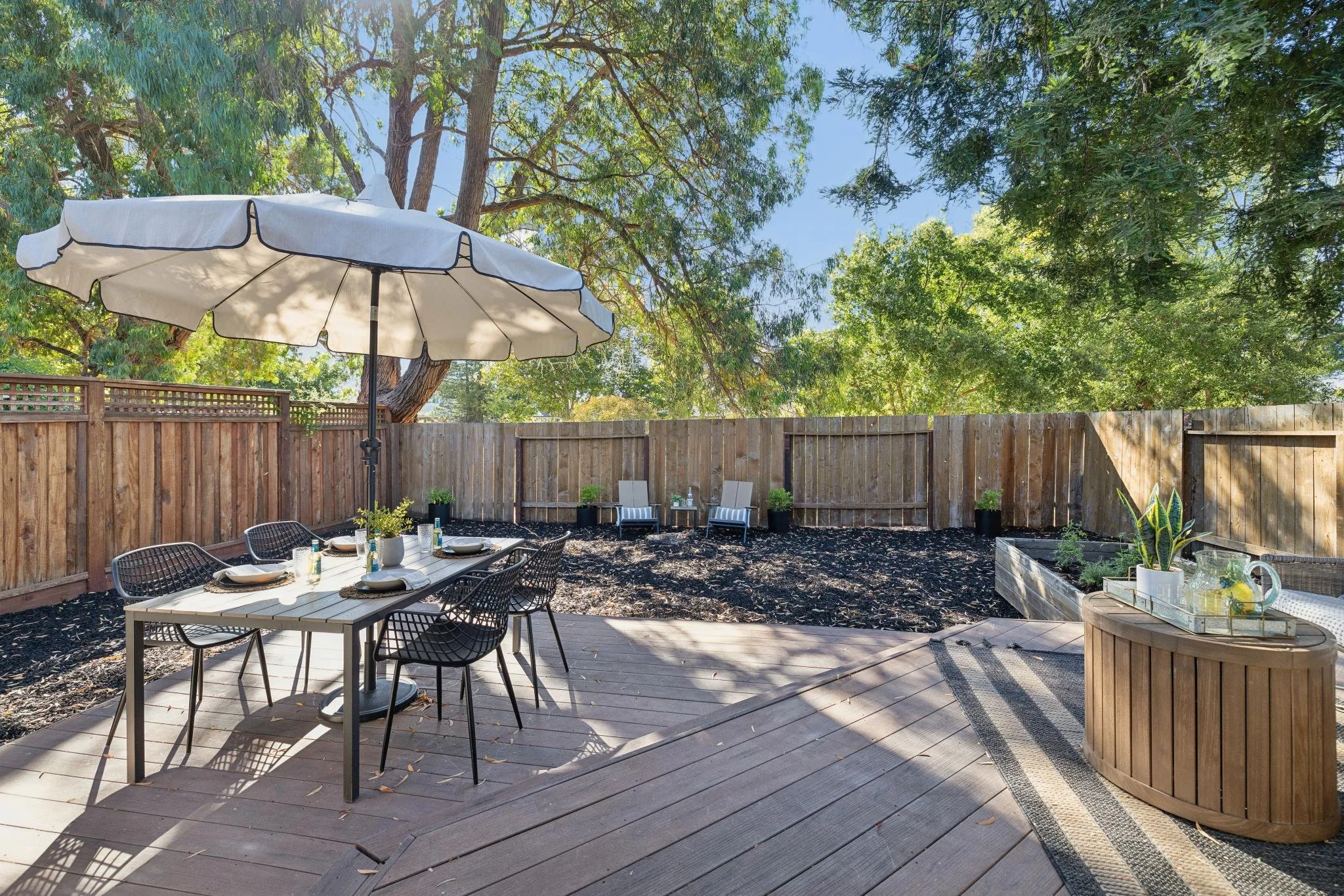 A backyard patio area with outdoor dining setup, including a table with chairs and an umbrella, surrounded by a wooden fence and lush green trees.