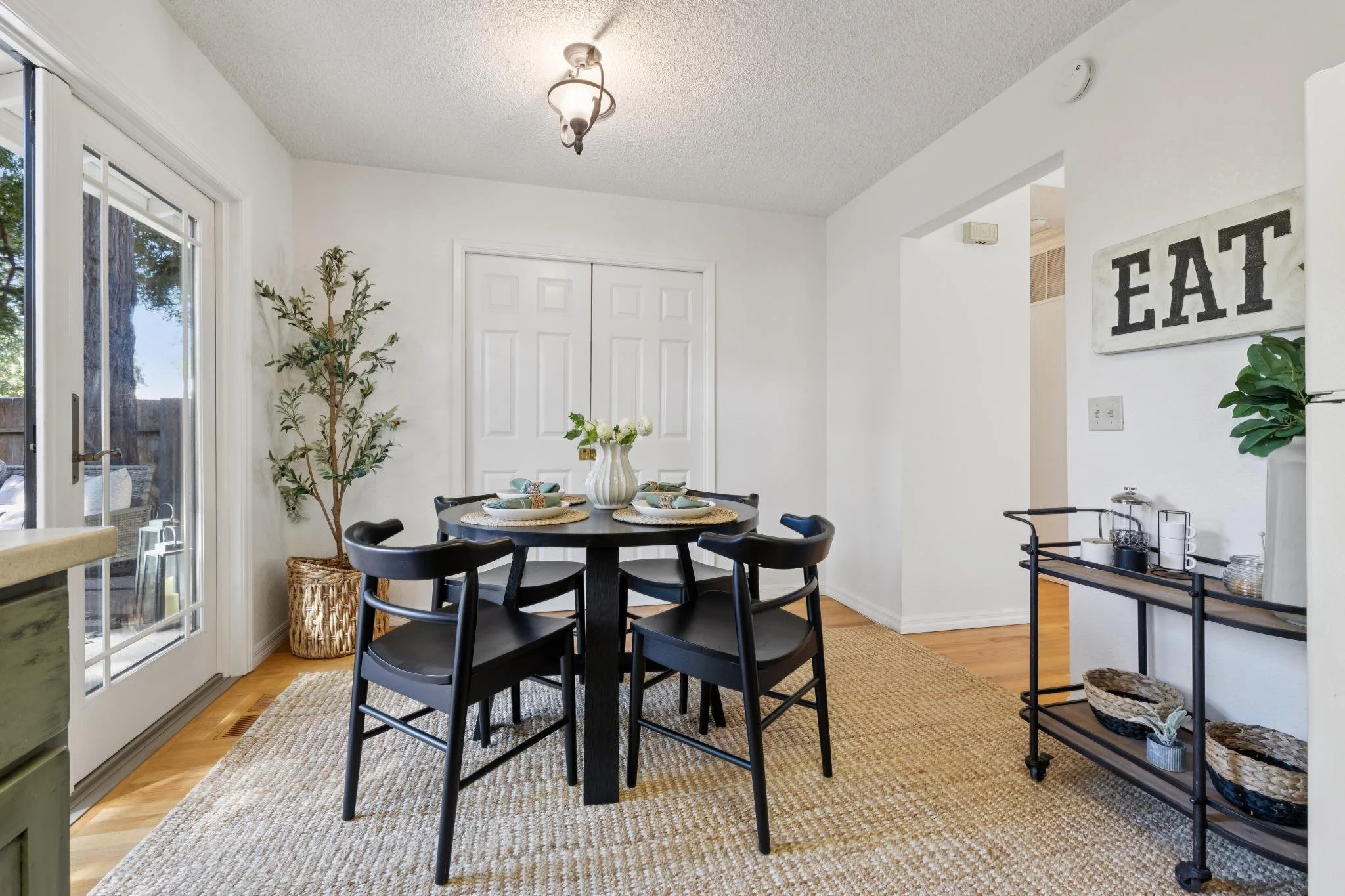 Dining area with a small round black table surrounded by six black chairs, a vase with white flowers on the table, a tall green plant in a woven basket, a cabinet with a tray of candles, and a sign that reads 'EAT' on the wall. The space has a light-