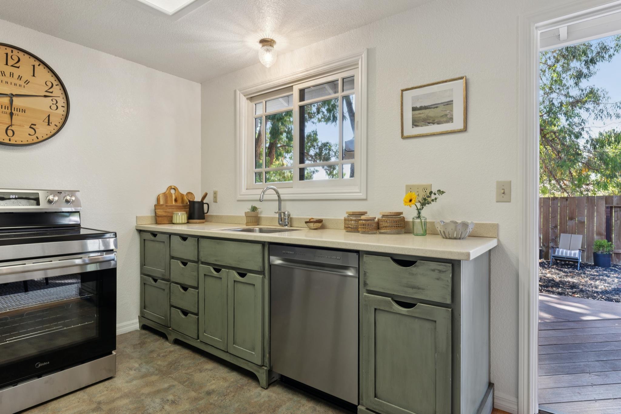A kitchen with a black stove, green cabinetry, a window above the sink, and a deck outside. Decor includes a sunflower in a vase, woven baskets, and wall art.