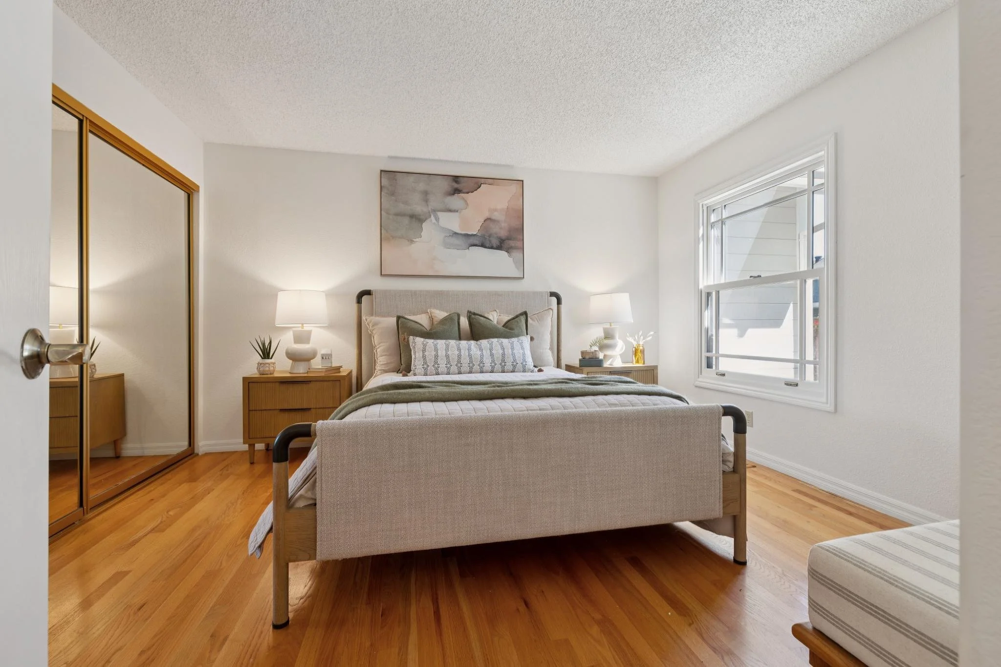 A bedroom with a bed, two nightstands with lamps, a large window, and a closet with mirrored sliding doors.