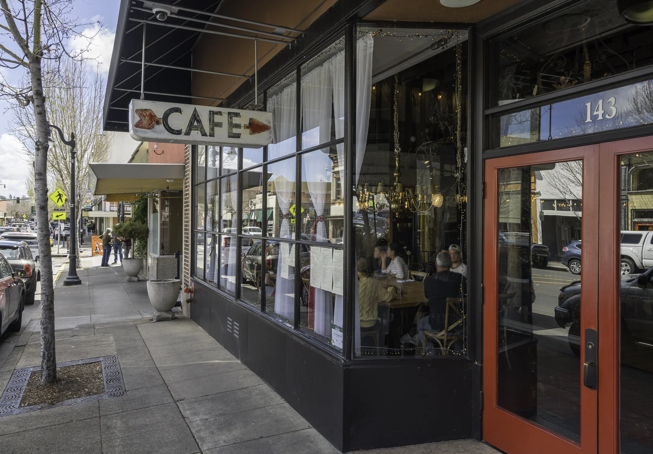 A storefront with large glass windows and red framing, displaying an interior with hanging chandeliers and string lights. There is a sign that reads 'CAFE' with red arrows pointing left and right. People are seated inside, and there are trees and par
