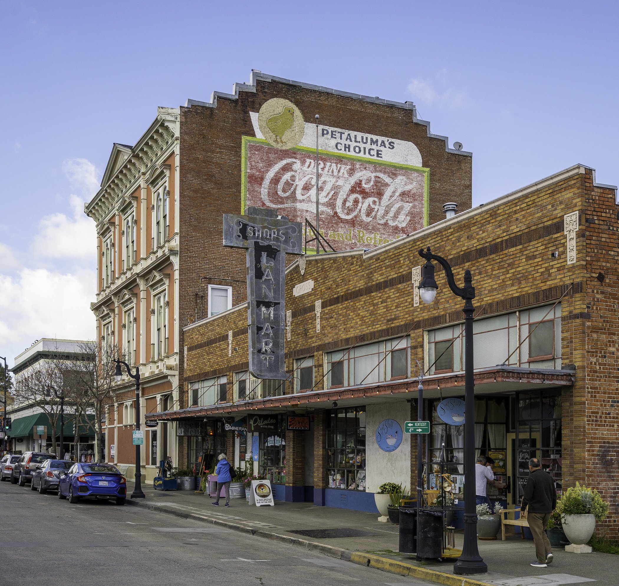 Street scene with historic brick buildings and storefronts, parked cars, street lamps, and people walking on the sidewalk, under a partly cloudy sky.