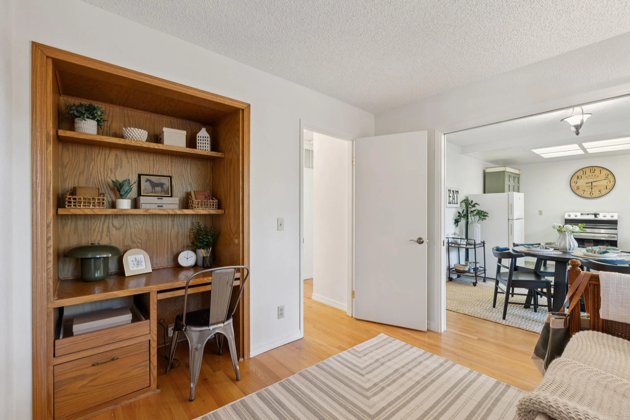 Living room with a wooden bookshelf, white wall, doorway leading to dining area, kitchen with clock, dining table with flowers, and hardwood floors.