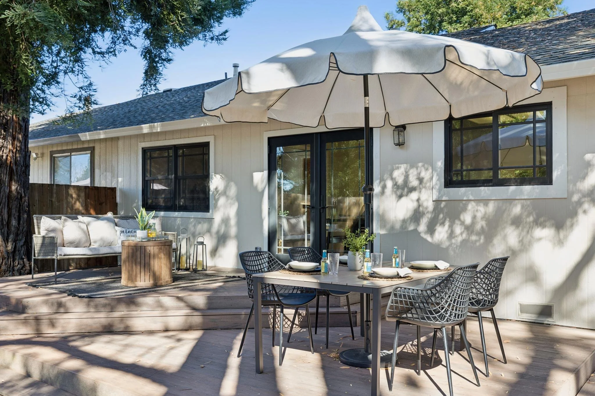 A backyard patio with a white house featuring large black-framed windows and a sliding door, a beige outdoor sofa with pillows, a round coffee table, a dining table with chairs and tableware, and a large white patio umbrella, surrounded by trees and 