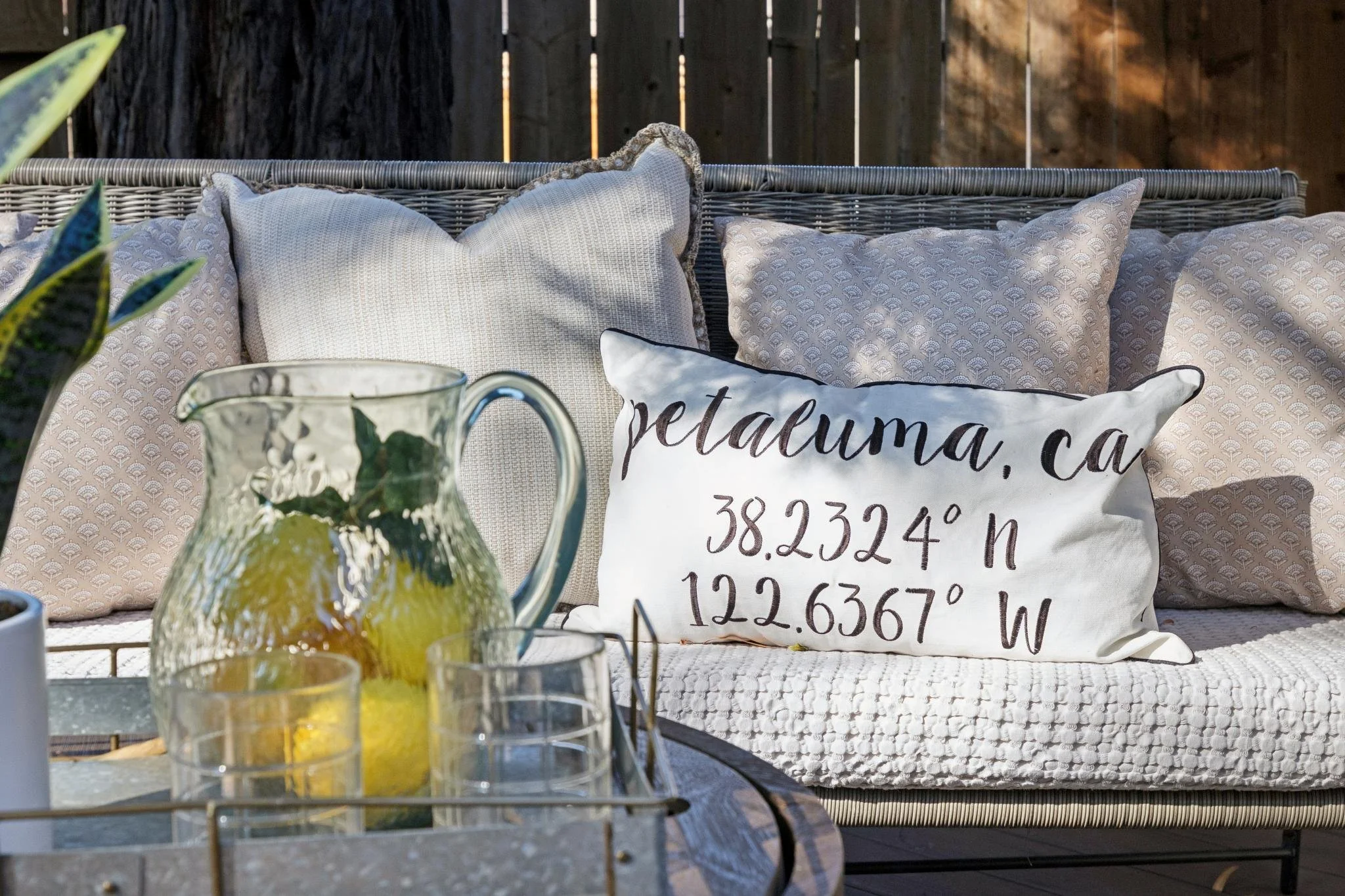 Outdoor patio area with a wicker bench decorated with multiple beige and patterned pillows. In the foreground, a glass pitcher filled with lemon slices and mint leaves, surrounded by small glasses, is placed on a table. A wooden fence is visible in t