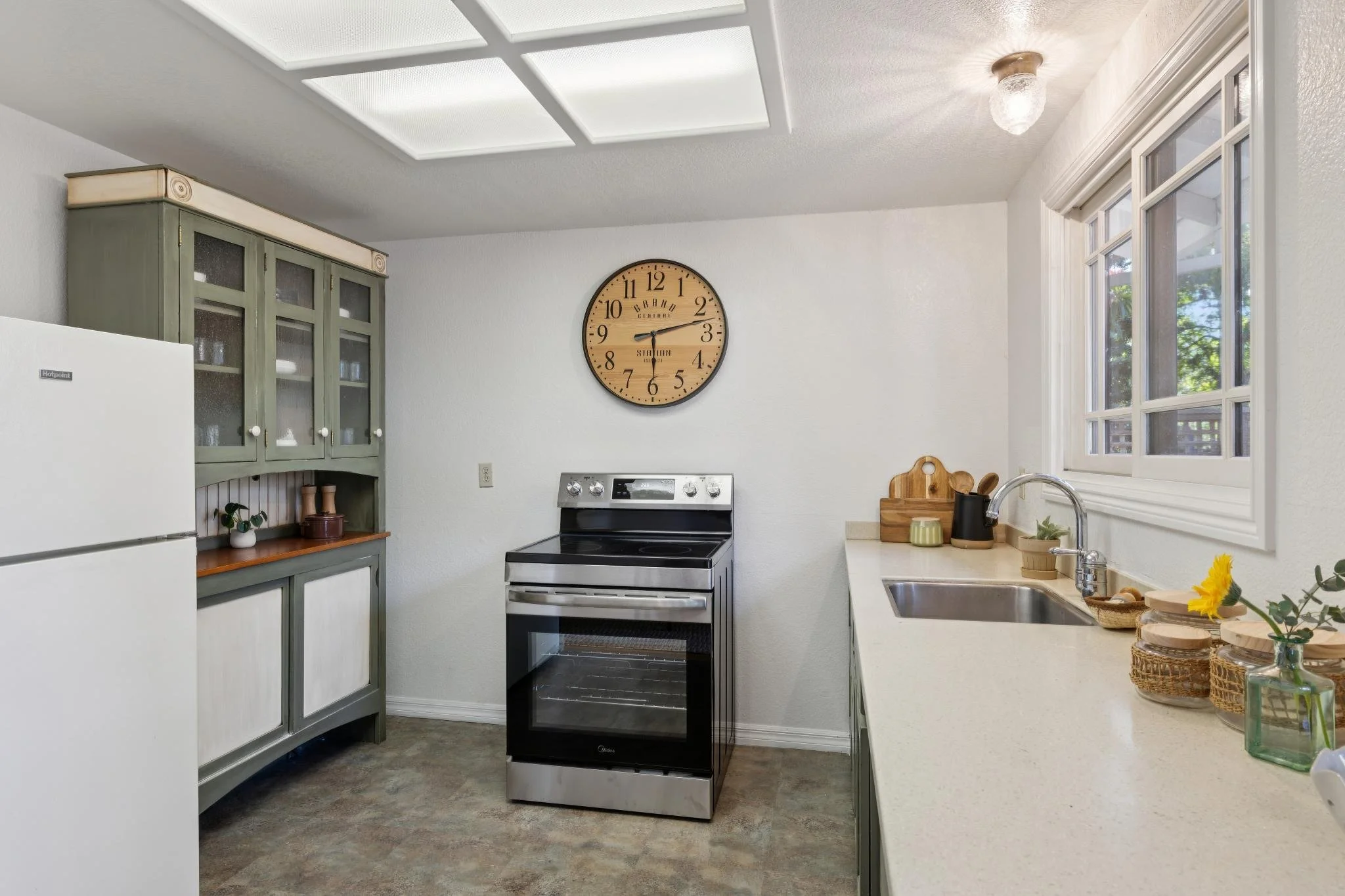 A kitchen with a white refrigerator, green cabinet with glass doors, black and stainless steel stove, countertop with a sink, window with white trim, wall clock, and various kitchen items and decor.
