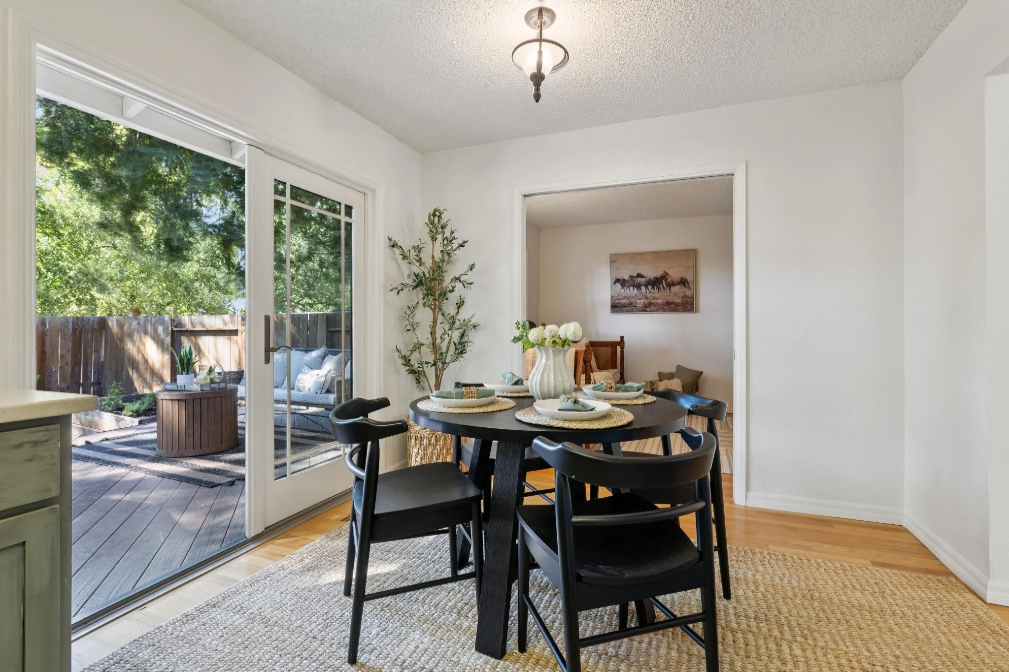 Dining area with black round table set with plates, napkins, and a white vase with flowers, overlooking a backyard patio through glass sliding doors.