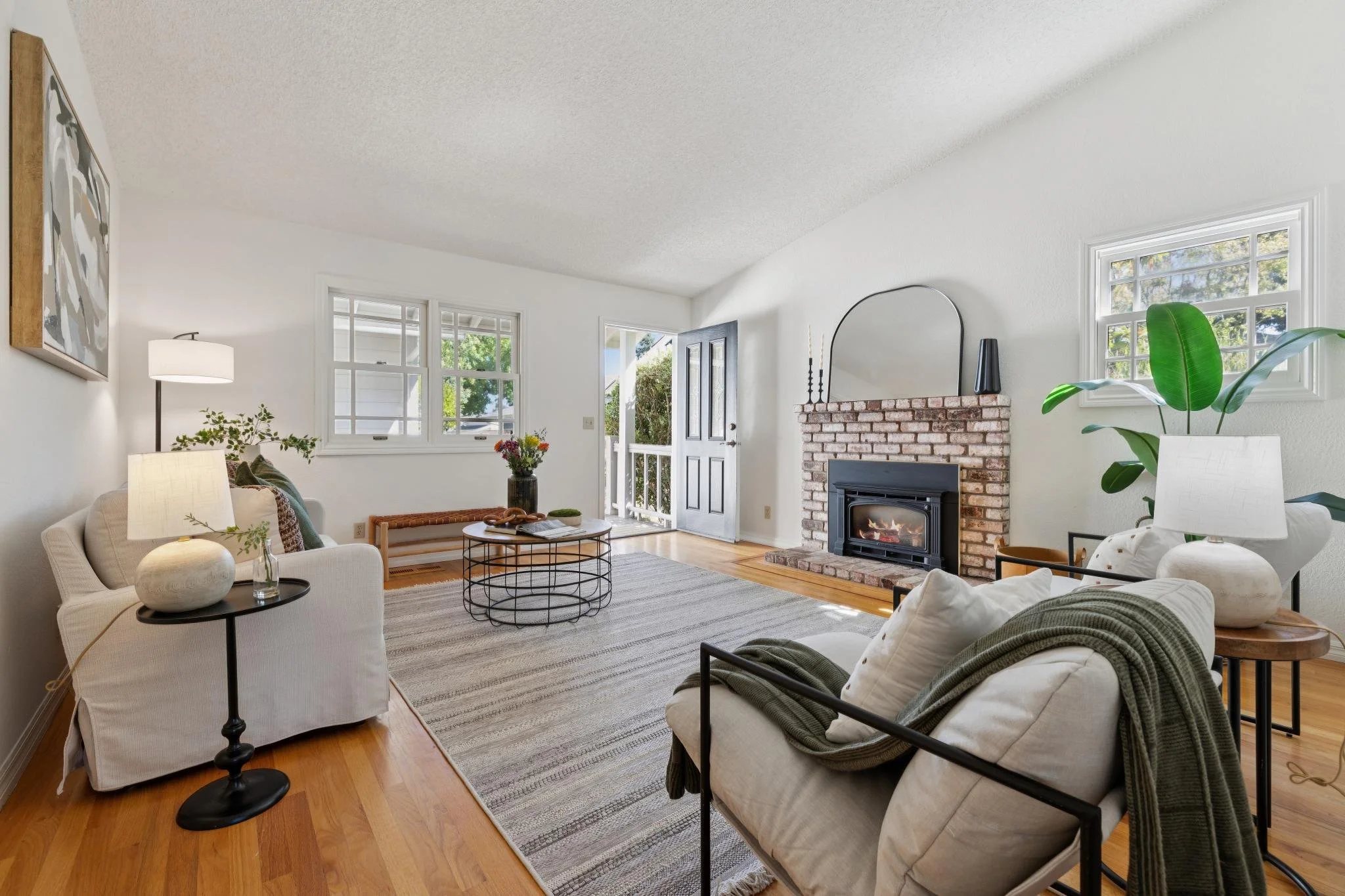 Bright living room with white walls, large windows, a brick fireplace, and modern furniture including white sofas and a black metal frame chair, accented with green plants and artwork.