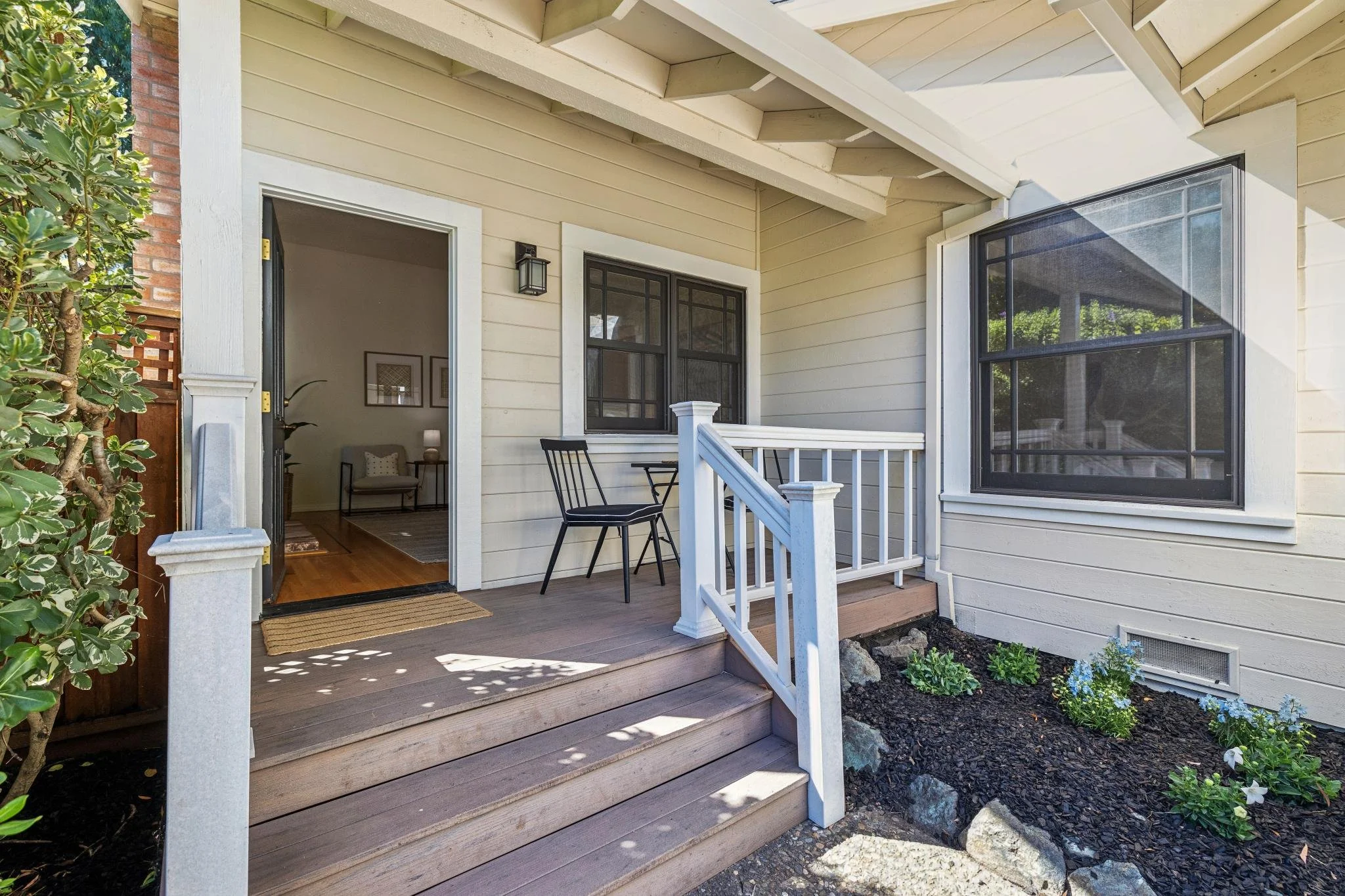 A small wooden front porch with stairs, black metal chairs, a flower bed with blue flowers, and a sliding glass door leading into a house.