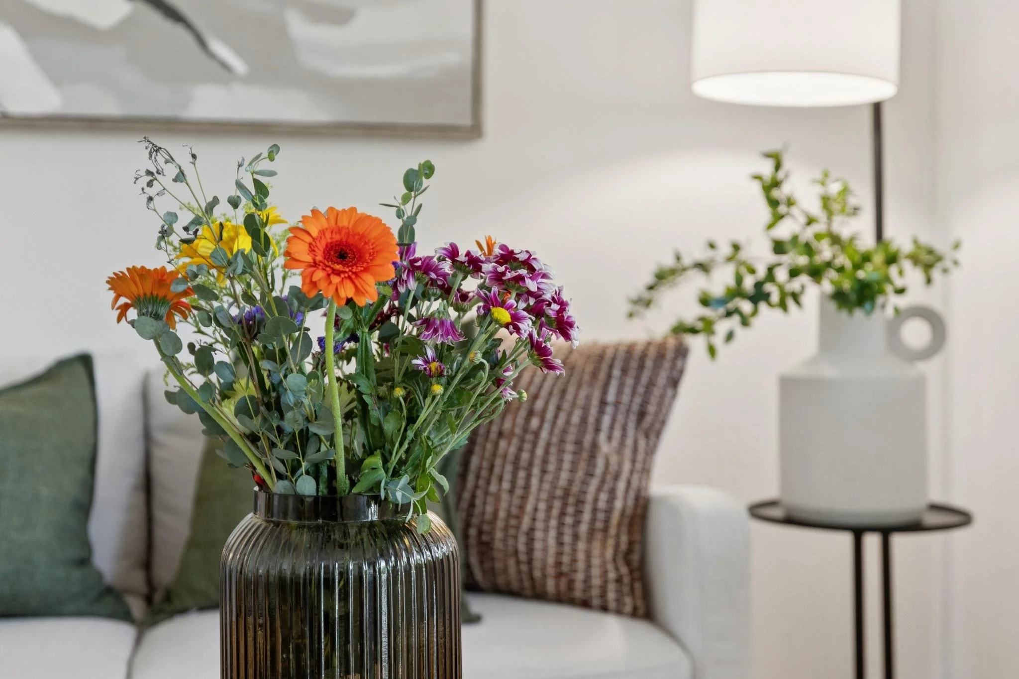 A bouquet of orange, pink, purple, and yellow flowers in a ribbed glass vase on a table in a cozy living room.