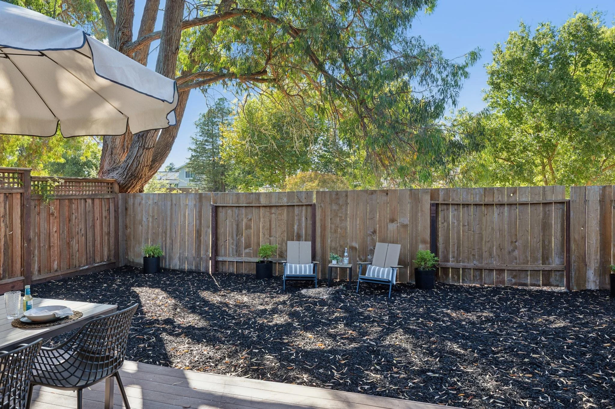 Backyard patio with outdoor furniture, potted plants, and a wooden fence. Large trees with green leaves overhead and a clear blue sky.
