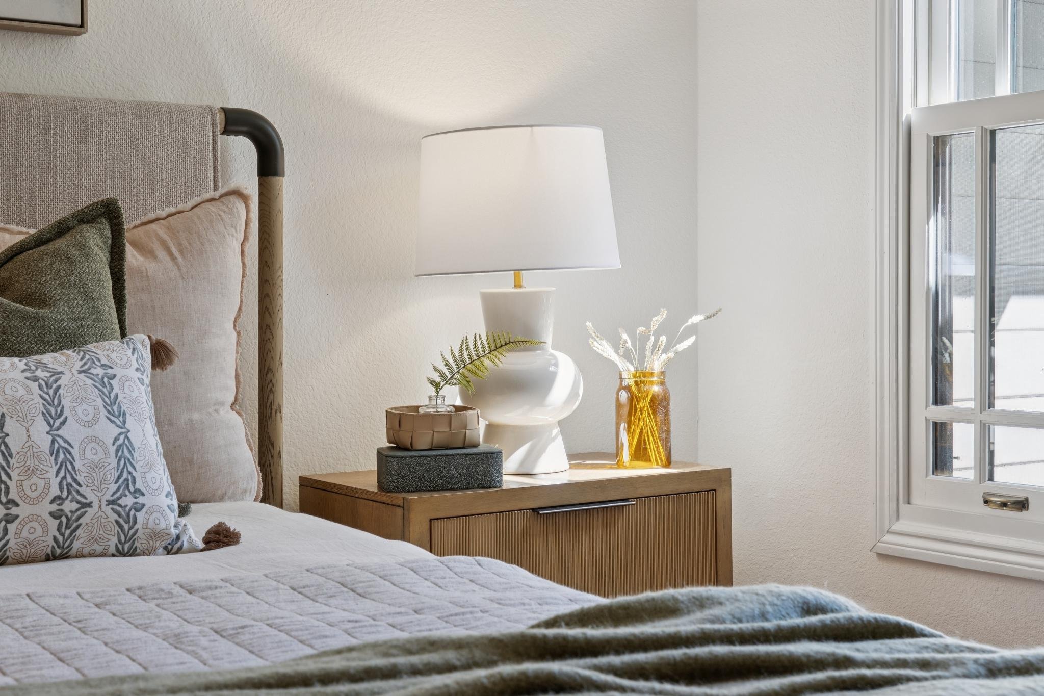 A cozy bedroom with a bed, pillows, a wooden nightstand, a white lamp, a yellow glass vase with dried flowers, near a window with white blinds, and light-colored walls.
