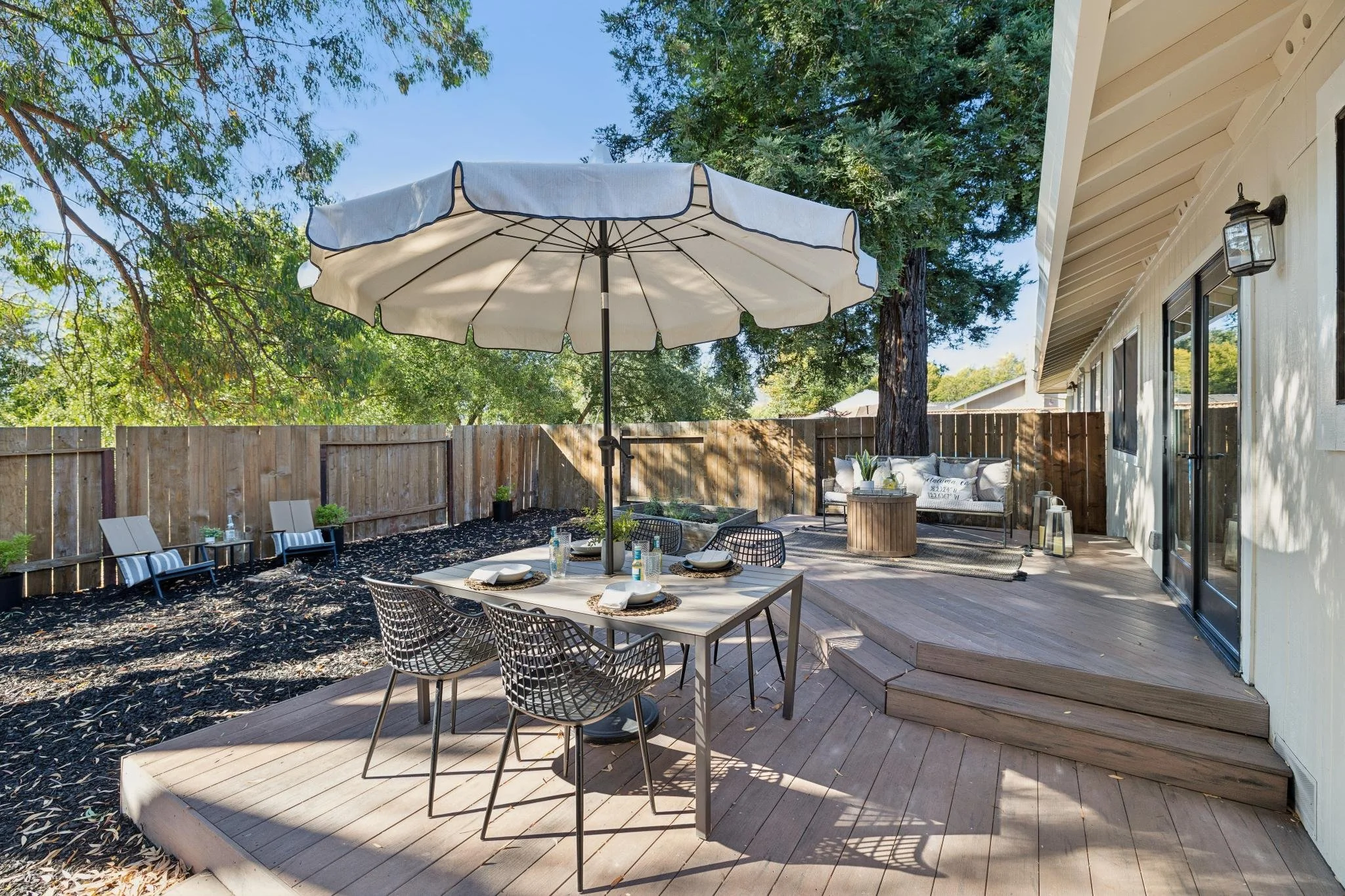 A backyard patio with a round table set for dining, an umbrella, outdoor seating, potted plants, and a wooden fence, adjacent to a house with large windows and an outdoor light.