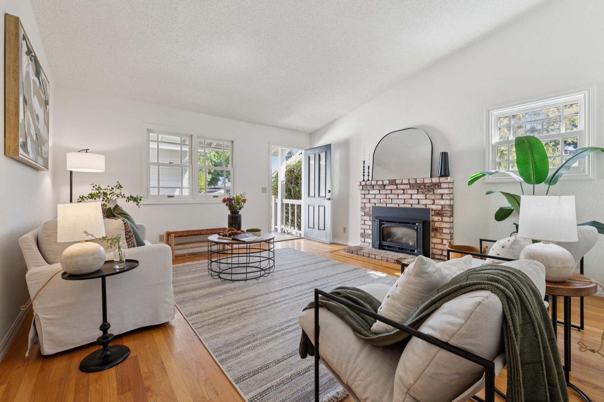 A living room with white walls, large windows, a brick fireplace, and contemporary furniture including a white sofa, armchair, and black metal accents. Decor features a large painting, potted plants, and a striped area rug.