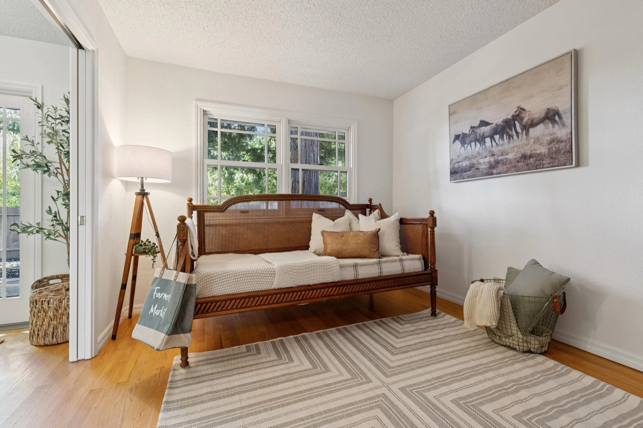 A cozy living room with a wooden settee, beige pillows, a floor lamp, and a painting of horses on the wall. Large windows show green trees outside.