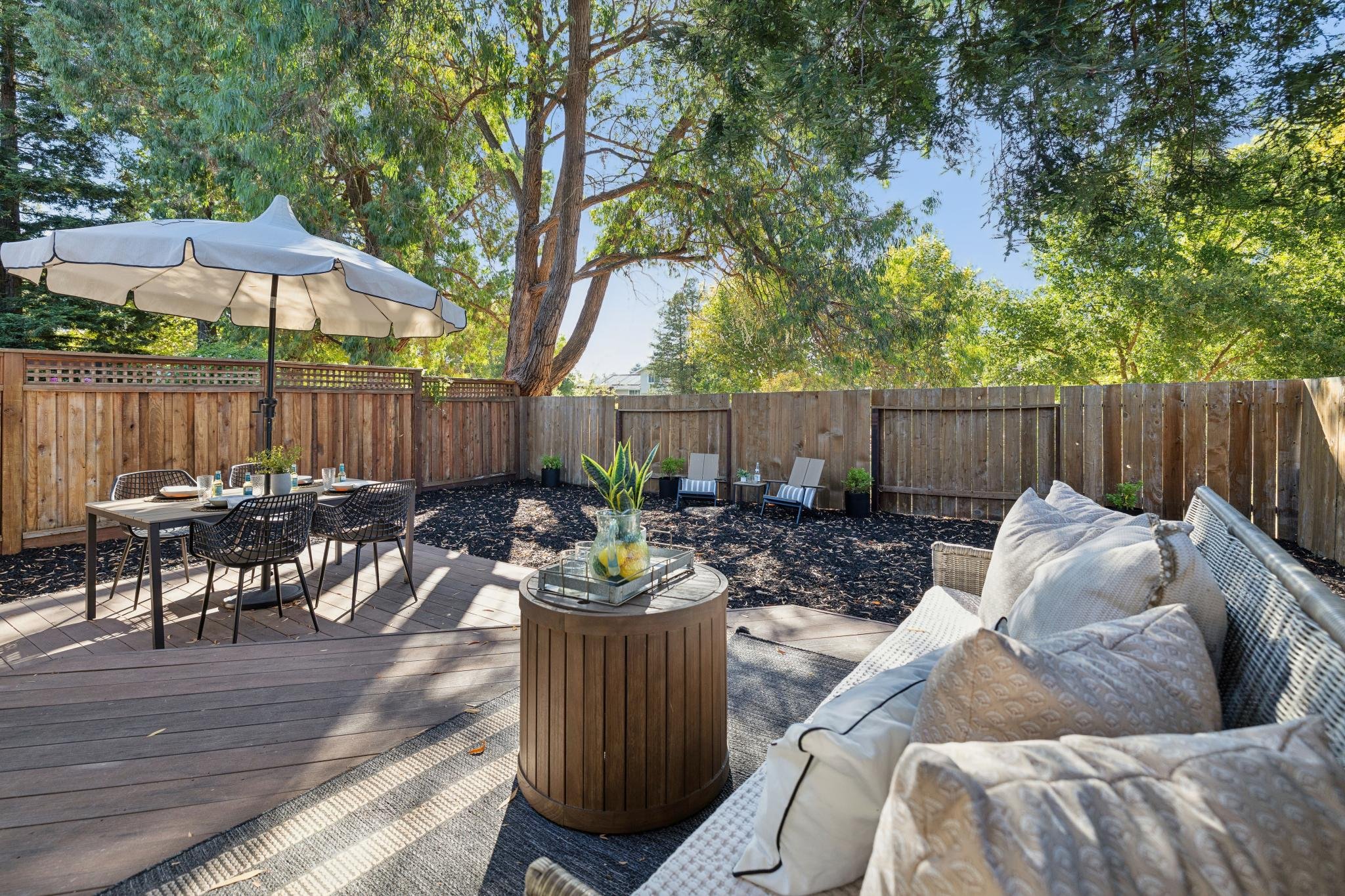 A backyard patio with outdoor furniture, a large umbrella, and surrounding trees and a wooden fence.