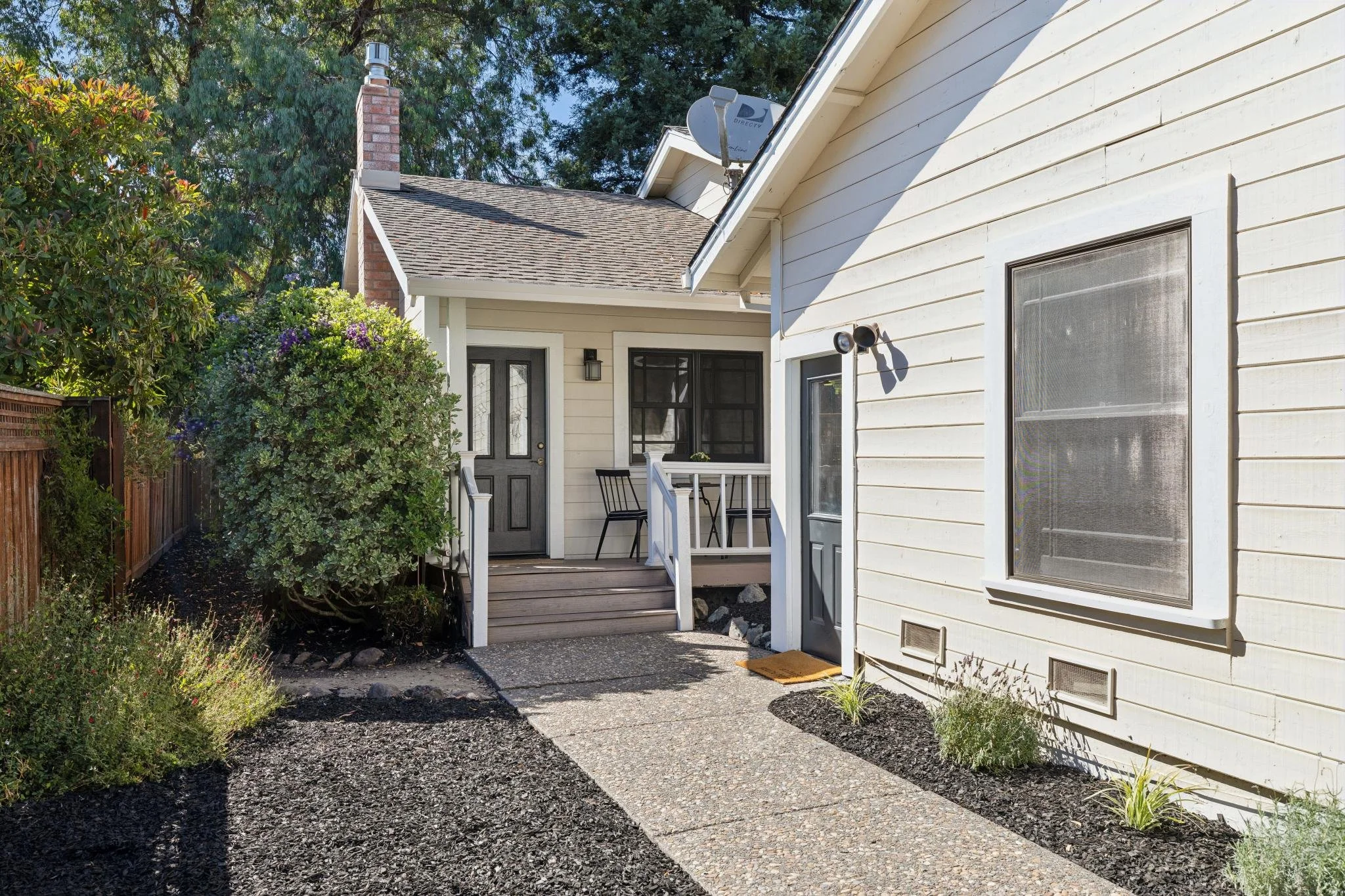 View of a residential backyard patio area with a white house exterior, black front door, small seating area with two chairs, and lush green shrubs and trees.