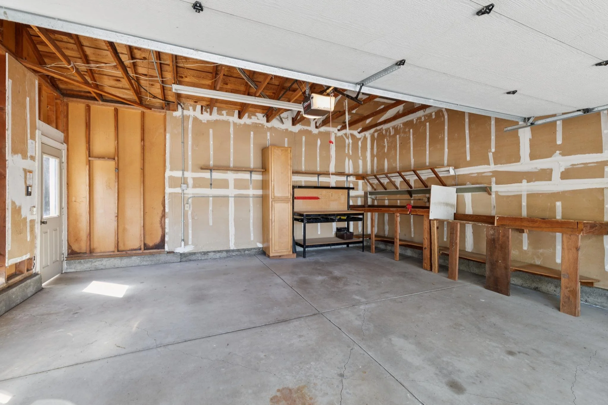 Empty garage with unfinished walls, exposed ceiling rafters, and some storage shelves and workbenches.