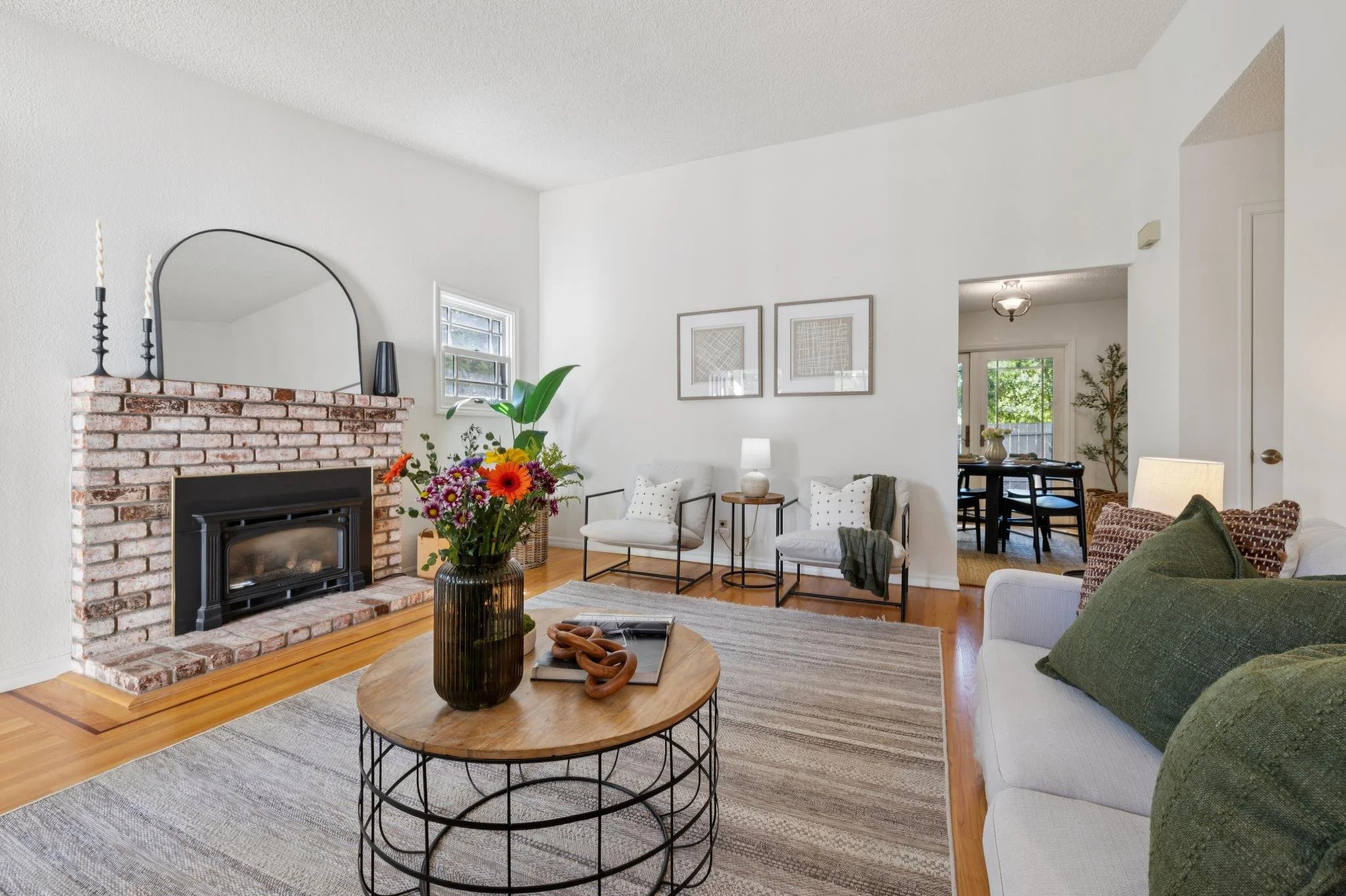 Living room with white walls, brick fireplace, round wooden coffee table with a black metal frame, colorful flower arrangement, green couch with dark green pillows, black metal framed chairs with white cushions, wall art, small wooden side table with