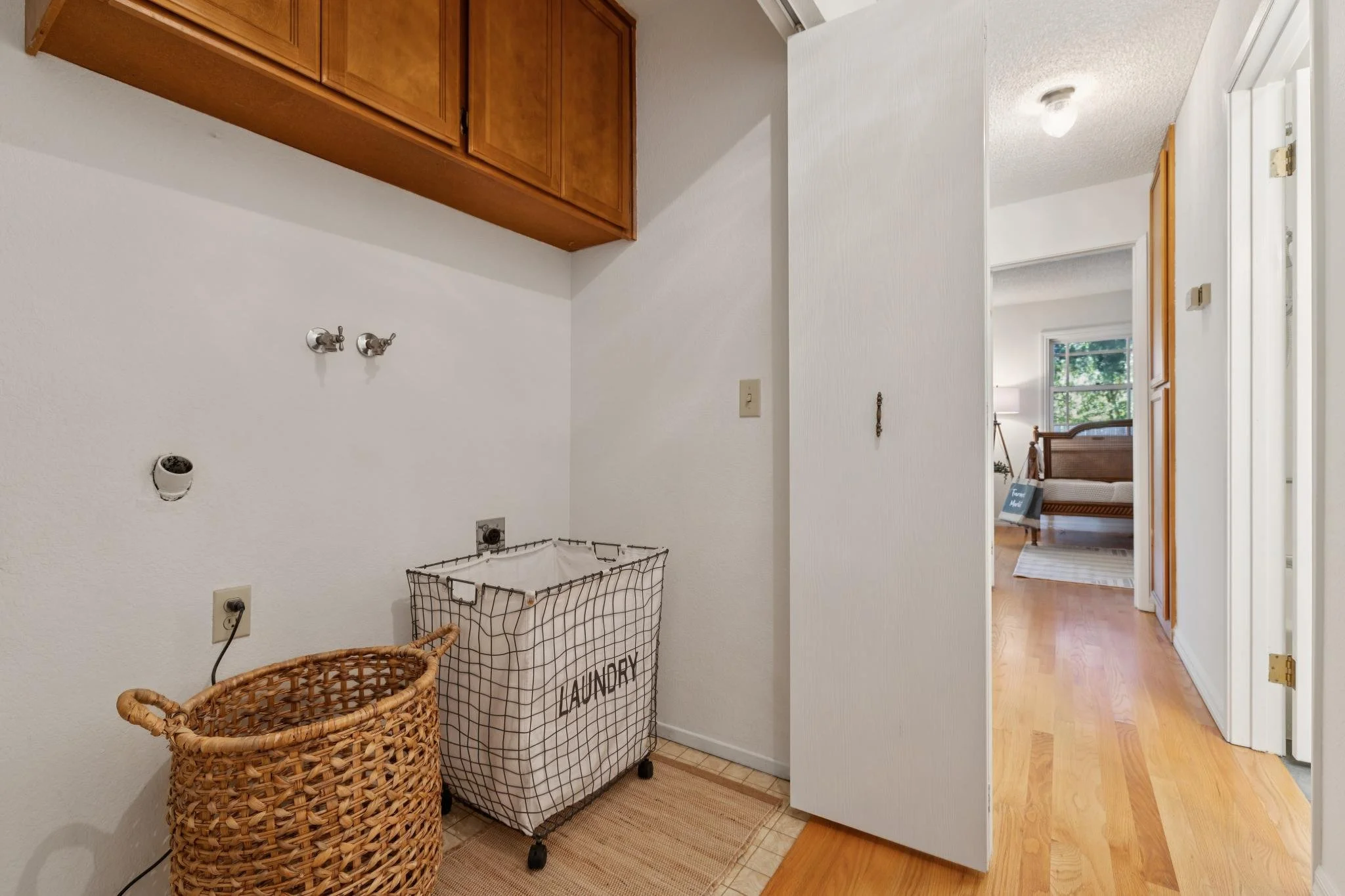 An empty laundry room with white walls, a wooden cabinet above, two water supply valves on the wall, a laundry basket on the floor, and a laundry bag with the word "LAUNDRY" printed on it; the room has a tiled floor and a view into a bright living ro