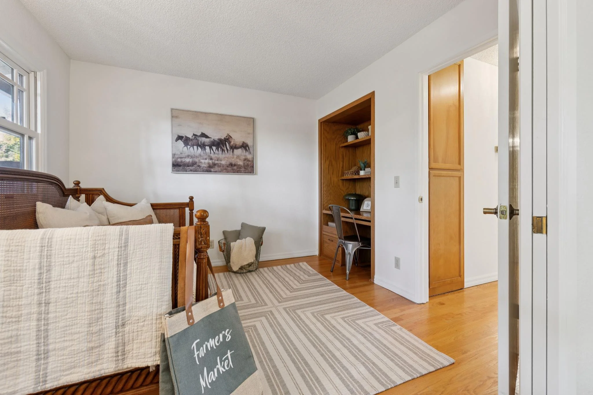 A cozy living room with a wooden bed frame and a white quilt, a framed picture of horses on the wall, a window with sunlight, a wooden bookshelf with decorative items and books, a metallic chair at a desk, a basket with blankets, and a striped rug on