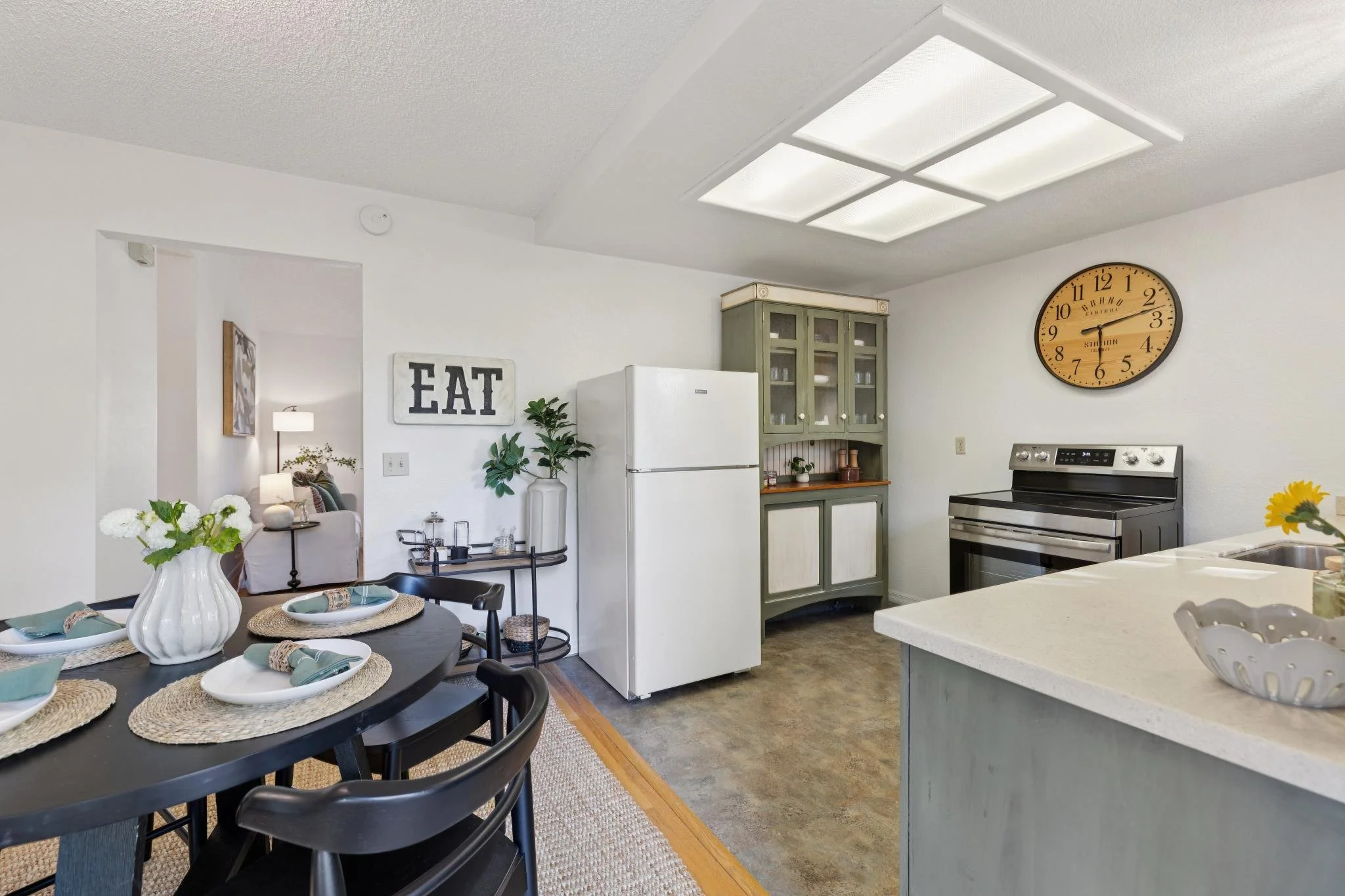 A kitchen and dining area with white walls, a black dining table set with white plates and green napkins, a vase with white flowers, a white refrigerator, and a green cabinet with glass doors. There is a large wall clock and a sitting area with a lam