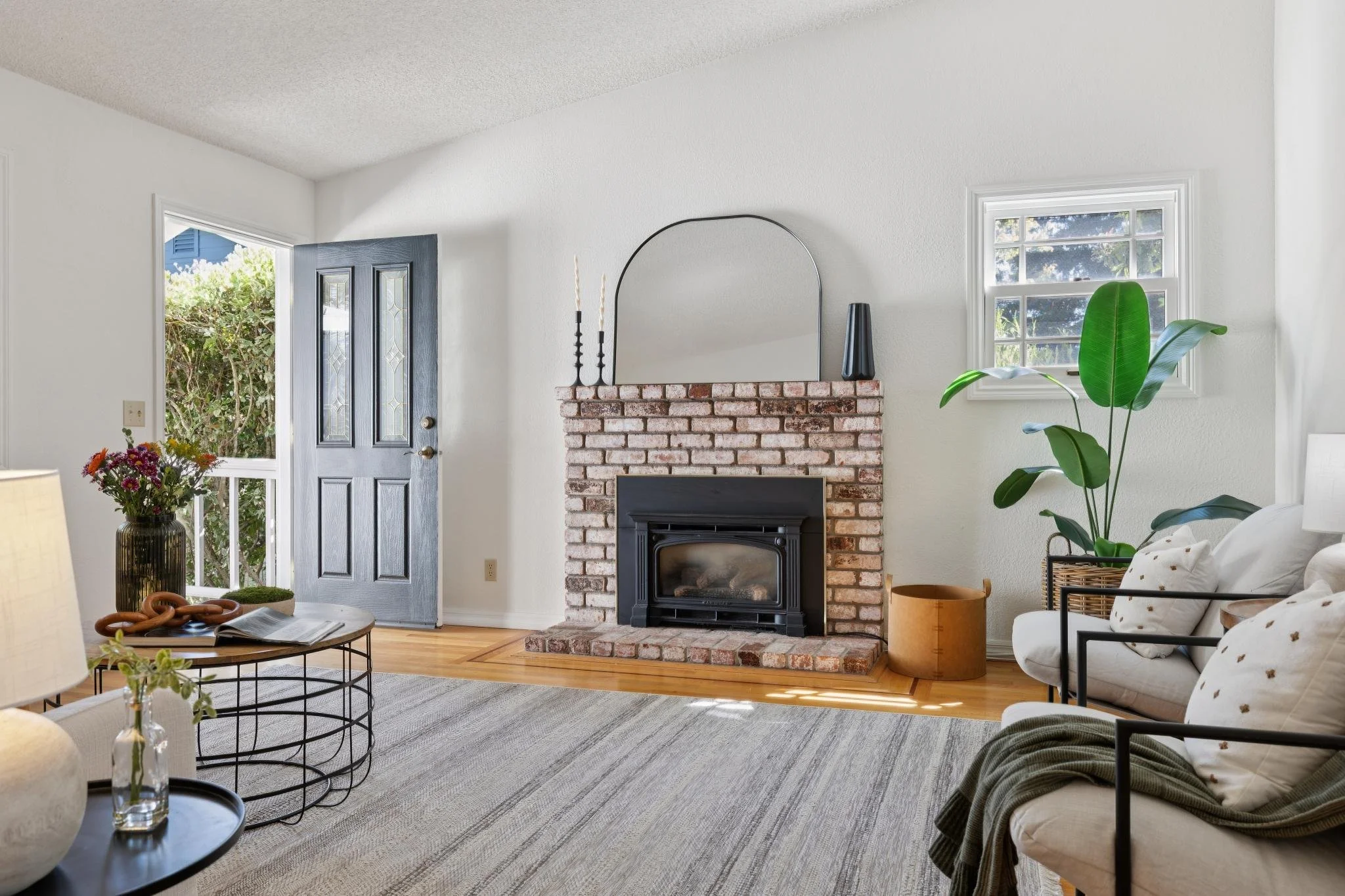 Living room with white walls, a brick fireplace, a black mirror above the fireplace, a large green plant, a window, a front door, a coffee table with a magazine, and a white sofa with pillows.