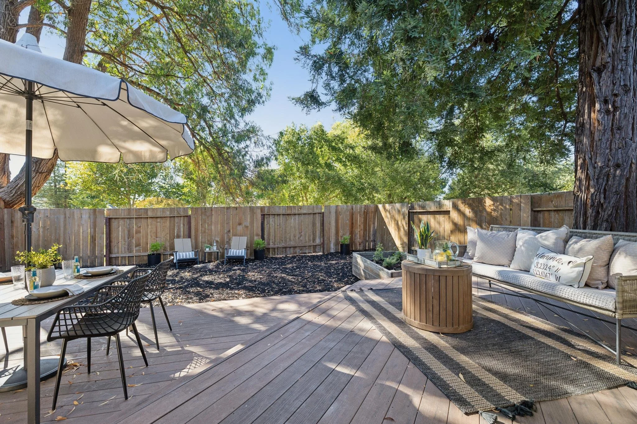An outdoor backyard patio featuring a dining table with chairs and place settings, a seating area with a cushioned sofa and pillows, a wooden fence, tall trees providing shade, a large umbrella, and various potted plants, all under a clear blue sky.