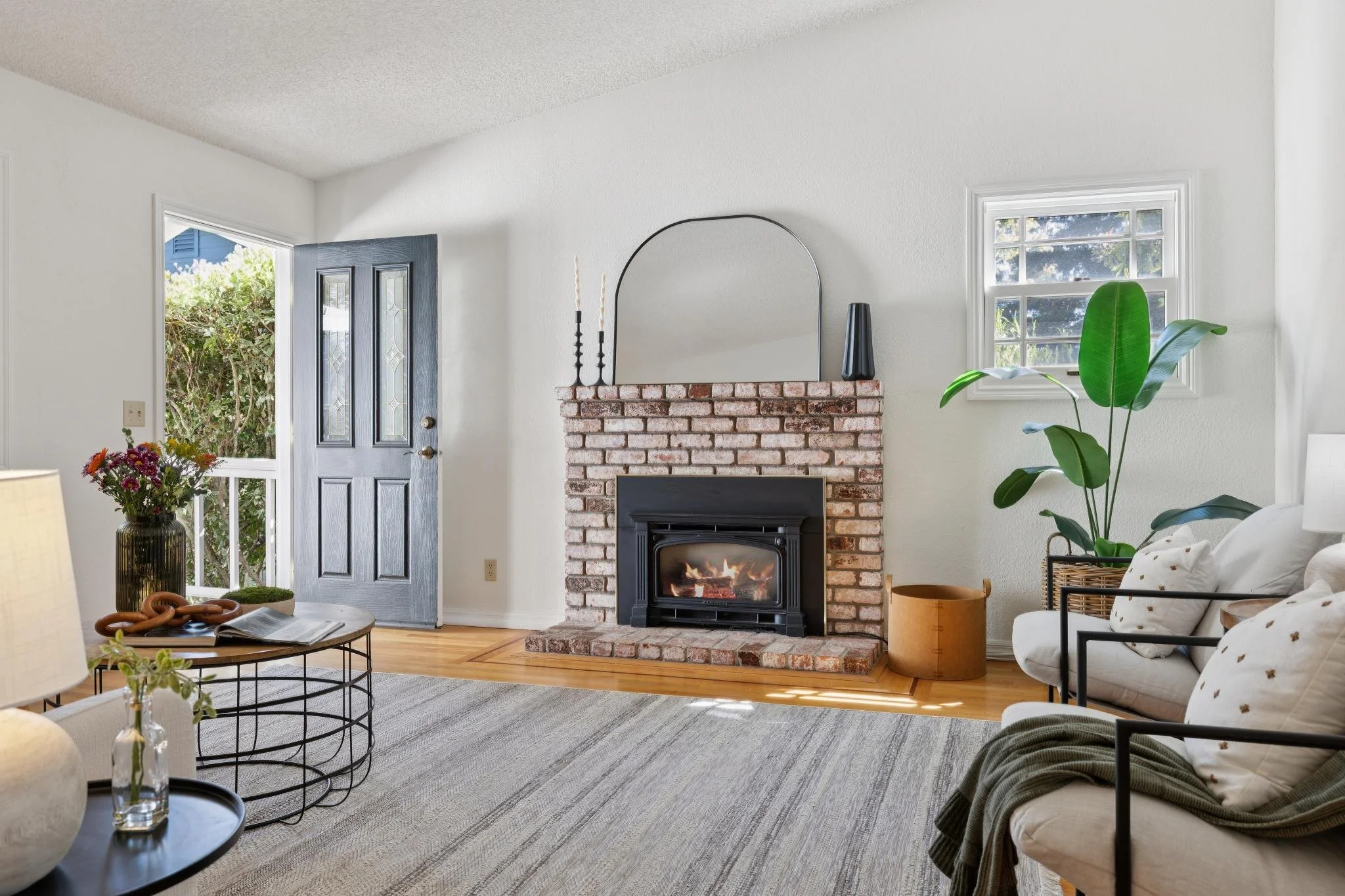 Interior of a living room with a fireplace, a front door, and windows letting in natural light. Decor includes plants, a mirror, and furniture.