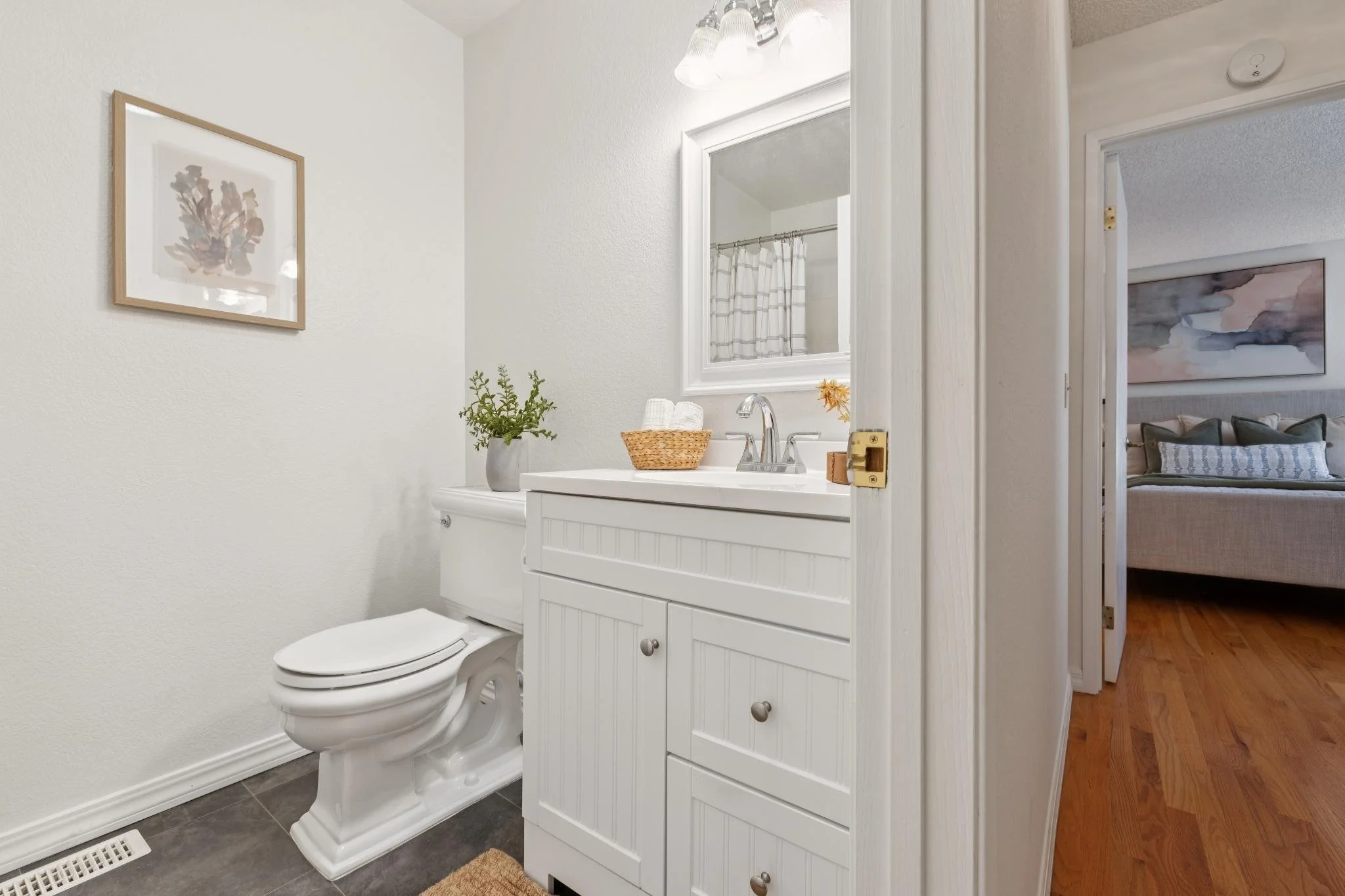 Bathroom with a white vanity, a mirror, a sink, a potted plant, and framed artwork on the wall. The toilet is next to the vanity, and a floor vent is visible on the dark tile floor. A doorway leads to a bedroom with a bed and wall art.