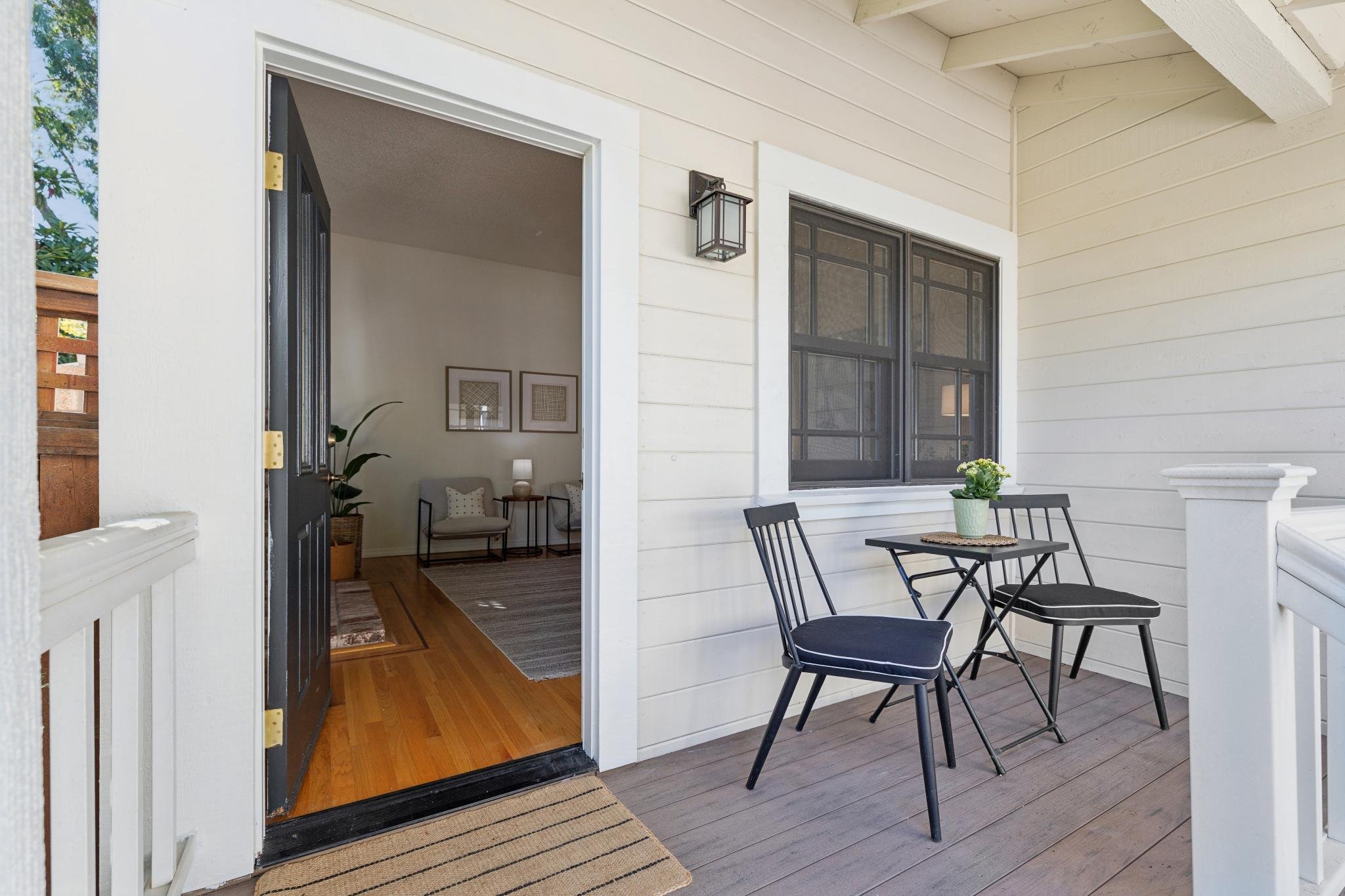 A porch area with a wooden floor and a table with four black chairs. There is a white railing, a potted plant on the table, and a window with a black frame. The interior of the house is visible through an open door, revealing a small sitting area wit