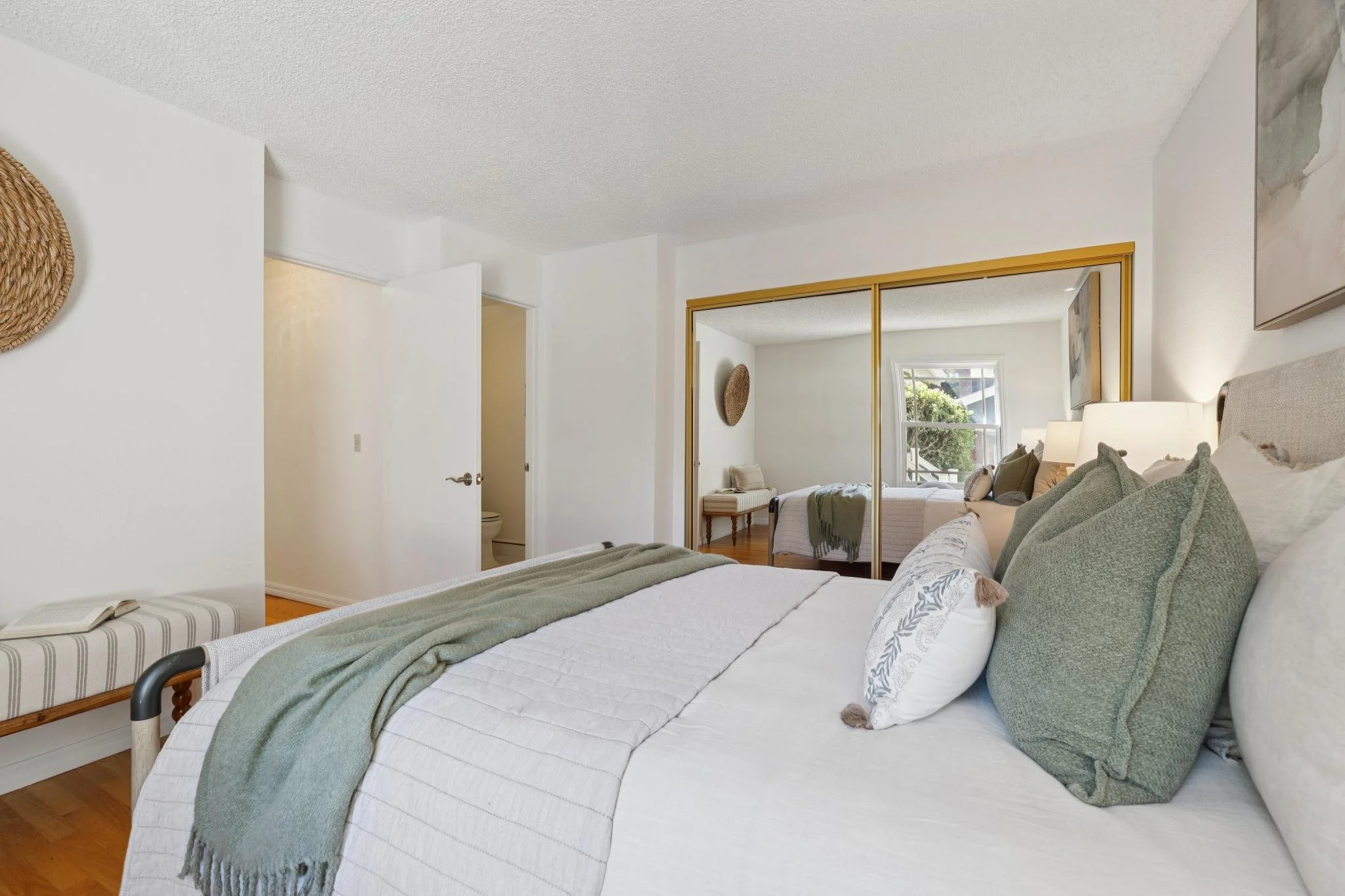 A cozy bedroom with a bed covered in white and green bedding and multiple pillows. There is a large window with a view of trees outside, mirrored closet doors, and decorative wall art. The room has wooden floors and white walls.