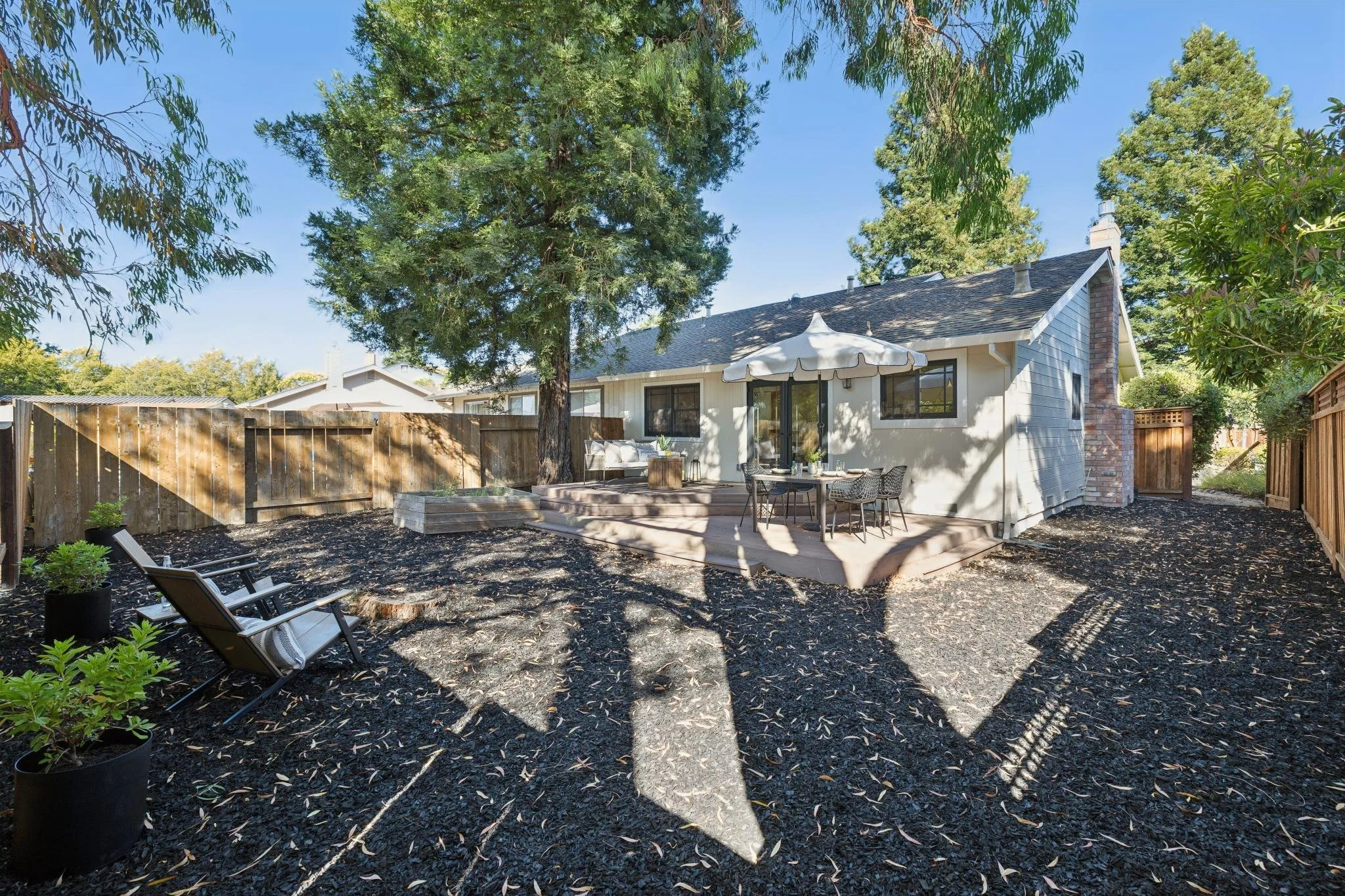 A backyard scene featuring a house with a deck, outdoor furniture, trees, and a clear blue sky.