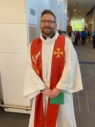 A man dressed in a white clerical robe with a red stole that has a gold cross and flame design, standing indoors at a church or conference center.