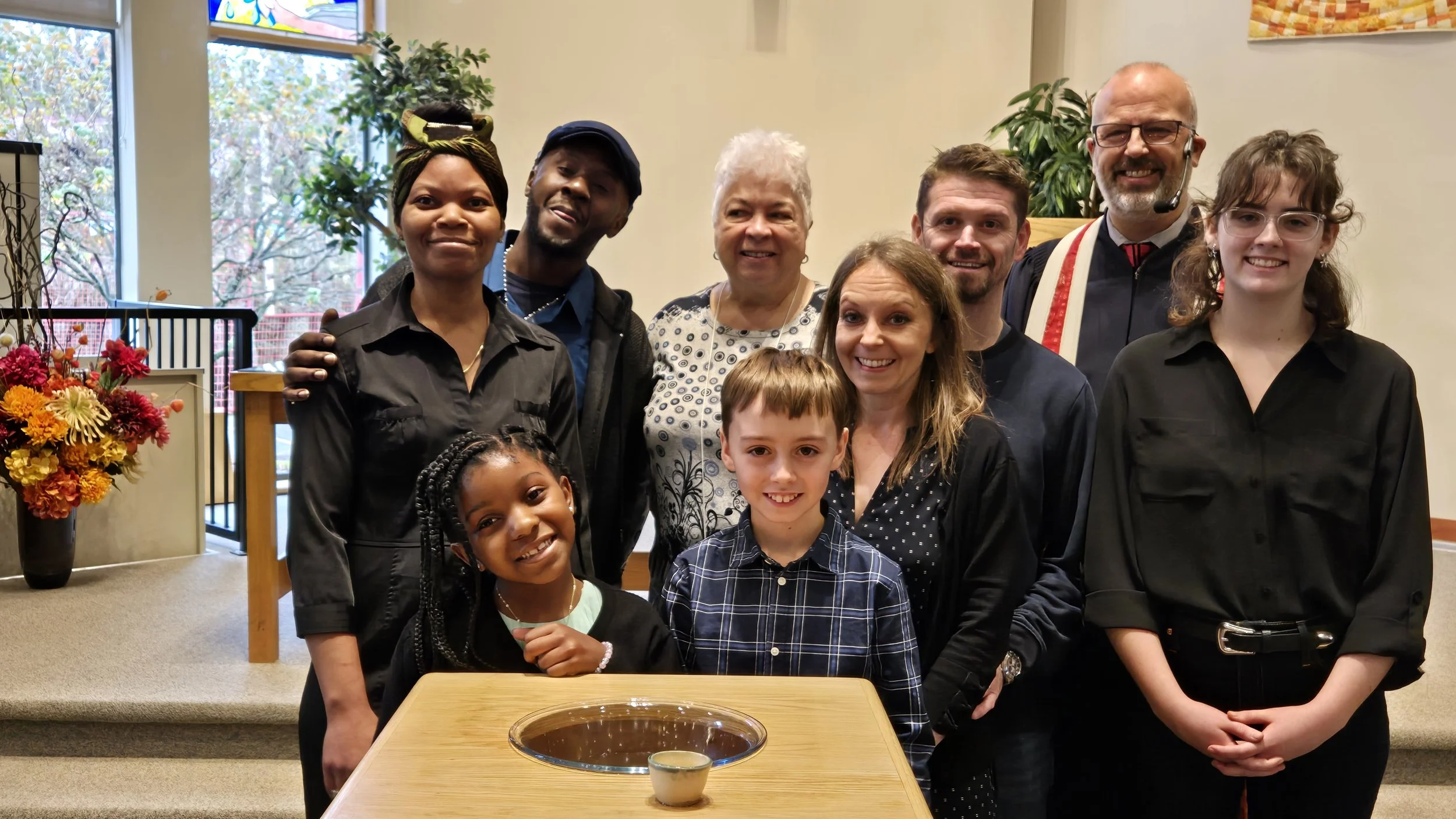 A diverse group of twelve people, including children and adults, posing together indoors near a table with a dessert in a bowl, with floral arrangements and windows in the background.
