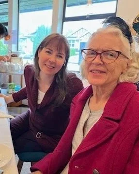 Two women sitting at a table in a bright room with large windows, smiling at the camera. One is elderly with glasses wearing a pink jacket, the other younger with long brown hair wearing a dark blazer.