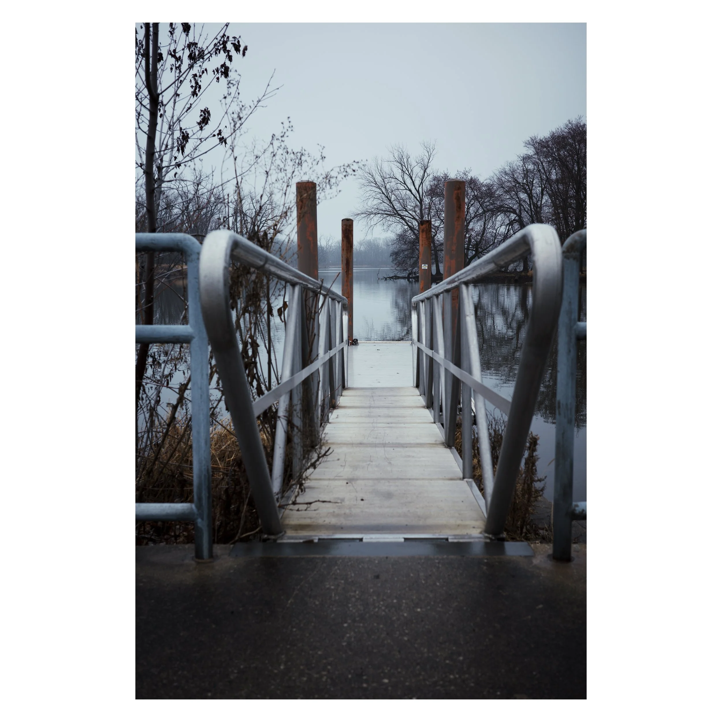 A metal dock or pier extends over a body of water with a river or lake in the background. The scene is outdoors during overcast weather with leafless trees and bushes on either side of the pier, suggesting late fall or winter. The water appears calm and reflective.