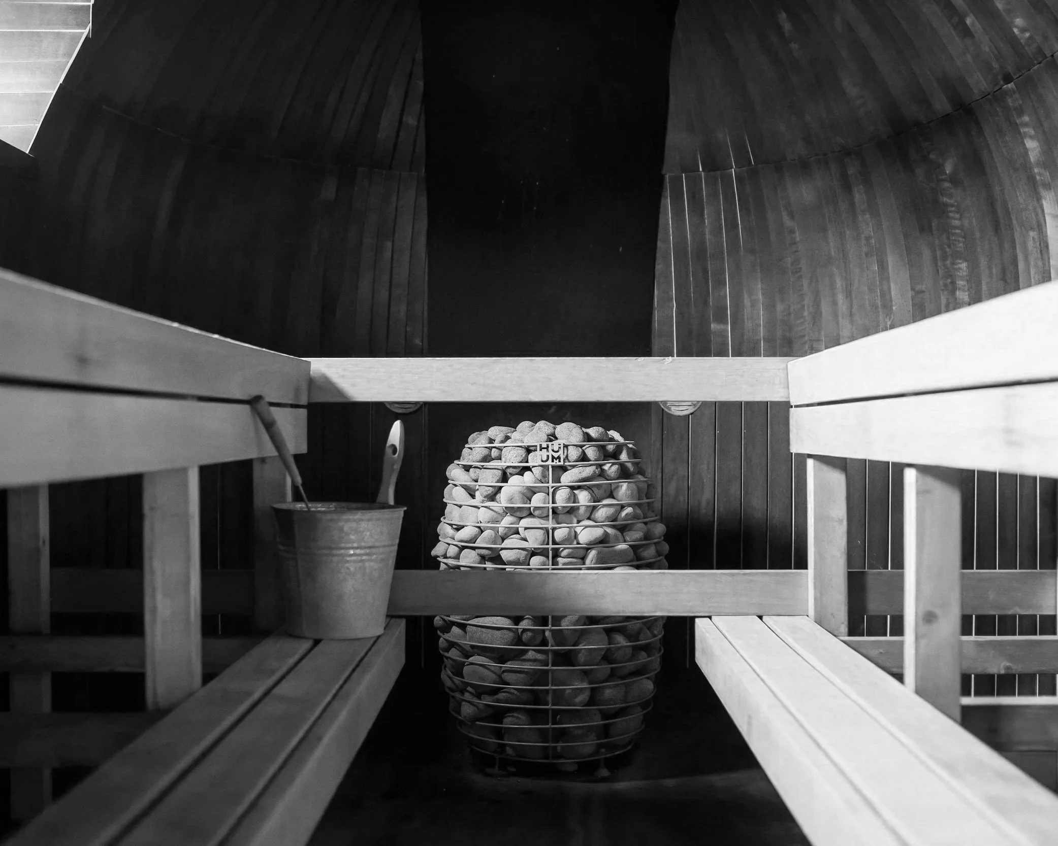 Black and white photograph of the interior of a sauna, with wooden benches and a central container filled with sauna stones. The image is taken from a high angle, looking down into the sauna.