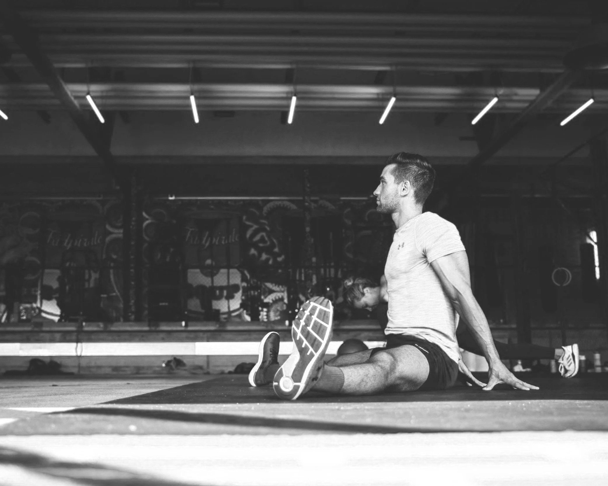 A man is stretching on the gym floor with his legs extended and hands on the ground, in a gym with workout equipment in the background.