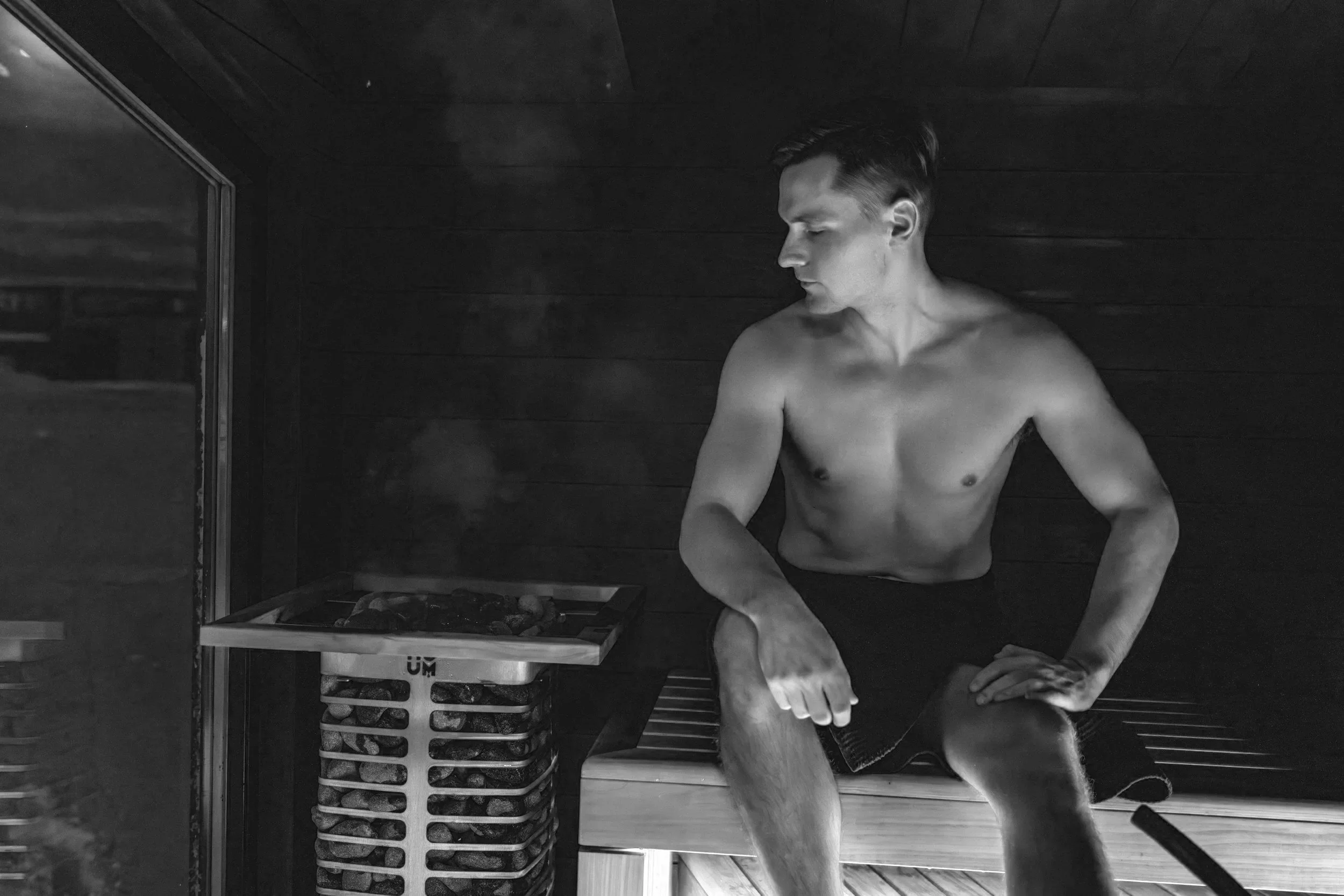 A shirtless man with short hair standing in a sauna, looking to the side, with wooden walls and floors around him.